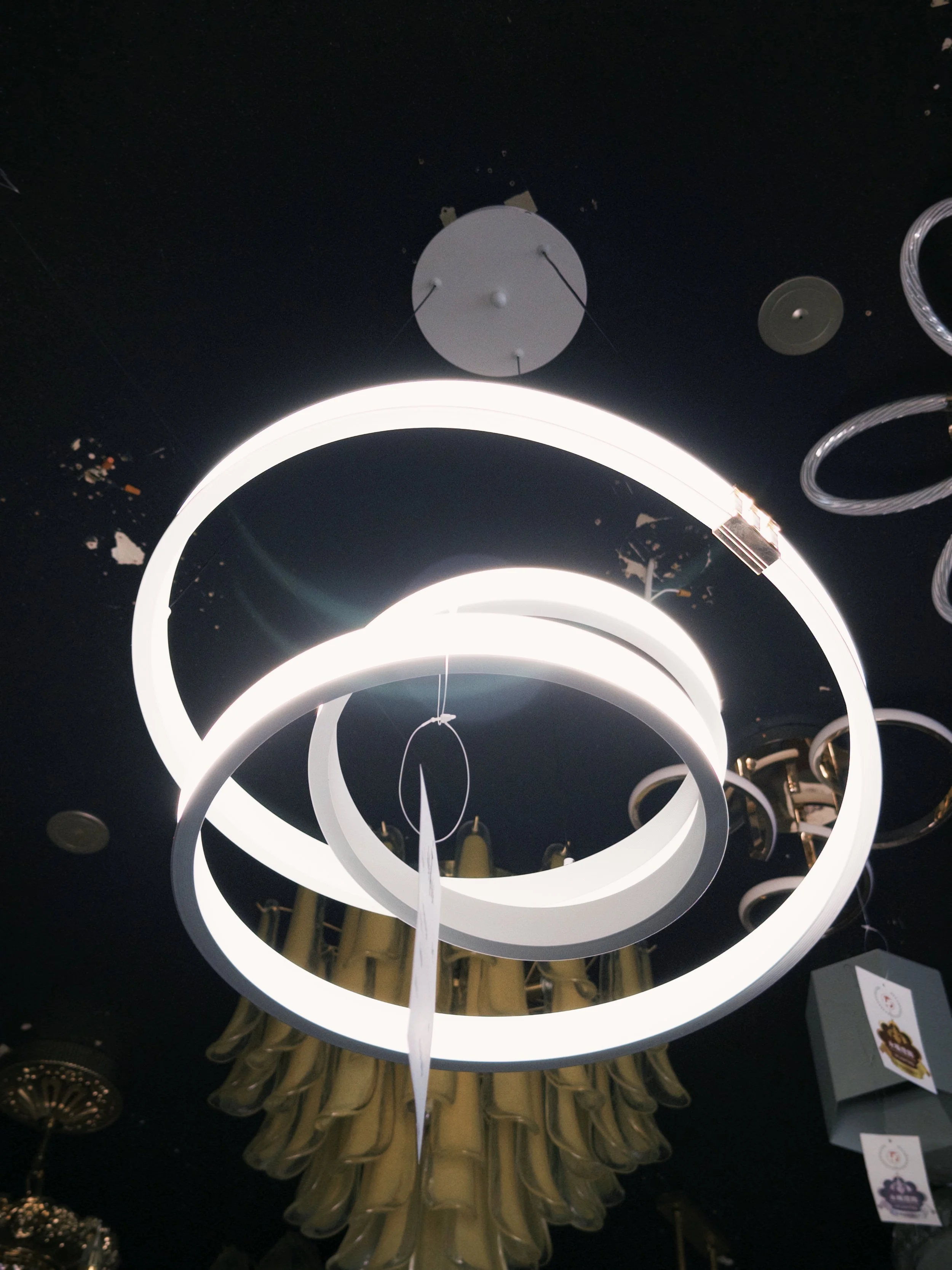 The image shows a modern ceiling light fixture with circular LED rings, hanging from a black ceiling. More bulbs and lighting elements are visible in the background.