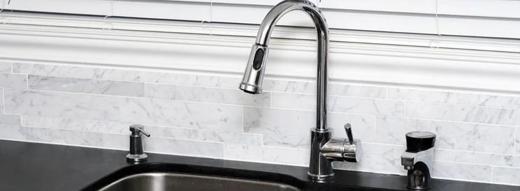 Kitchen sink with a chrome faucet and soap dispenser against a white marble backsplash and horizontal blinds.