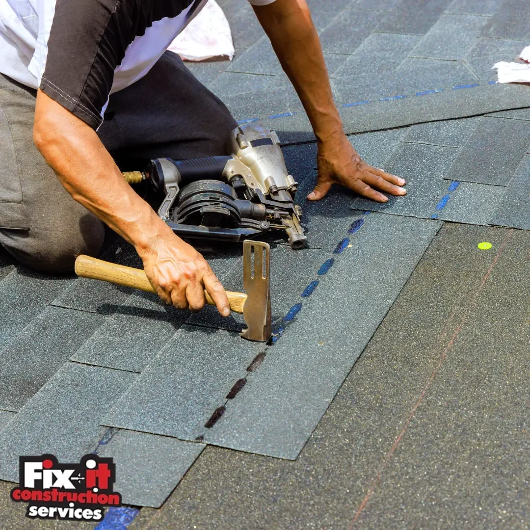 Close-up of a roofer kneeling on a gray asphalt shingle roof using a pneumatic nail gun and holding a roofing hatchet while installing new shingles along a chalk-line course, representing storm shingle