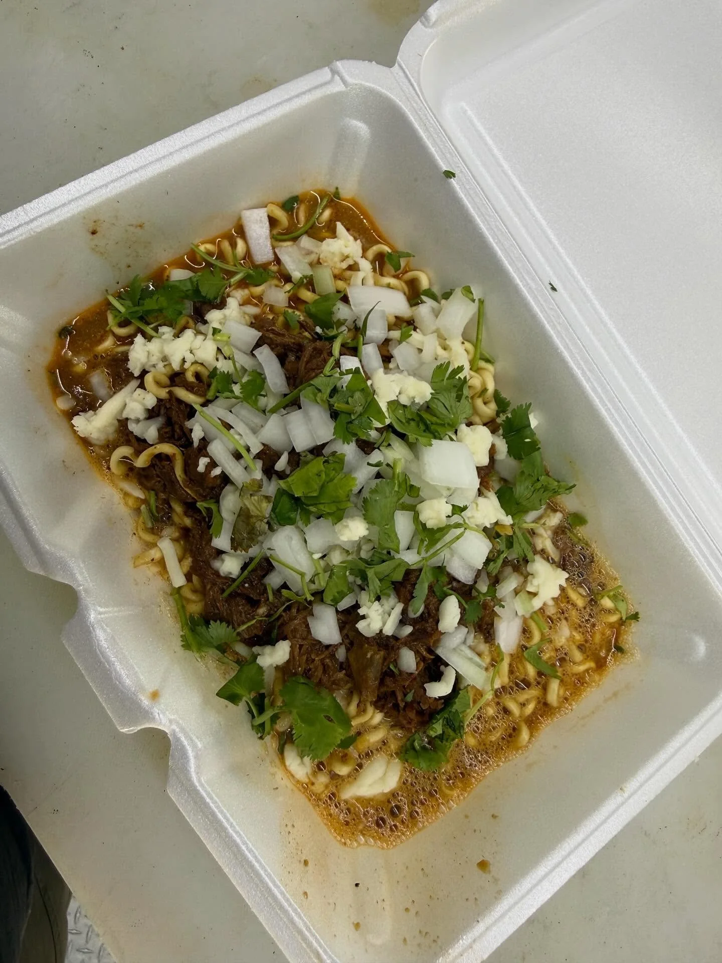 To-go container of ramen noodles topped with chopped onions, shredded beef, shredded cheese, and cilantro.