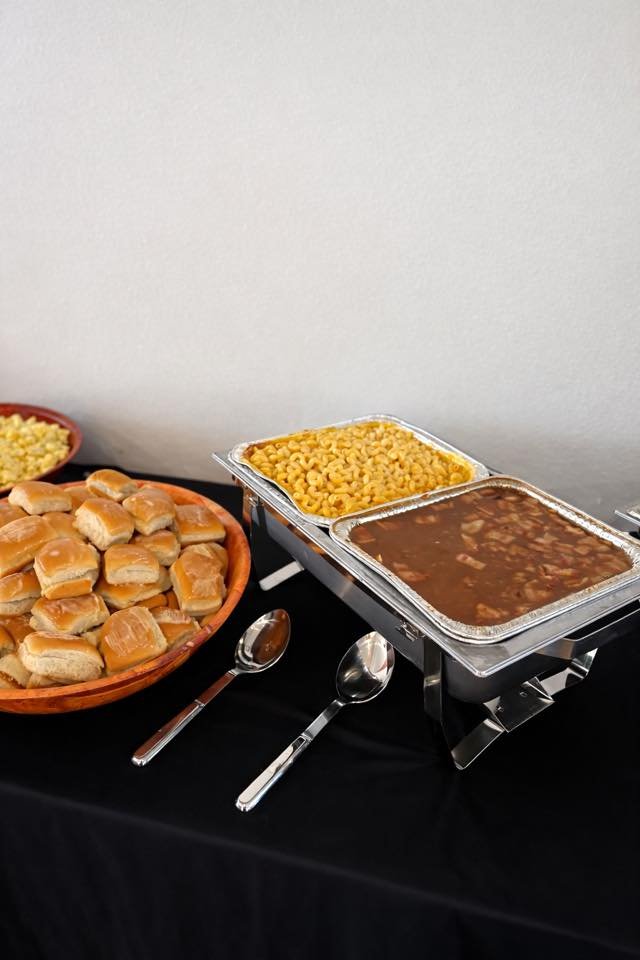 Buffet table with slider buns, macaroni and cheese, beef stew, and baked beans, with serving spoons and a black tablecloth