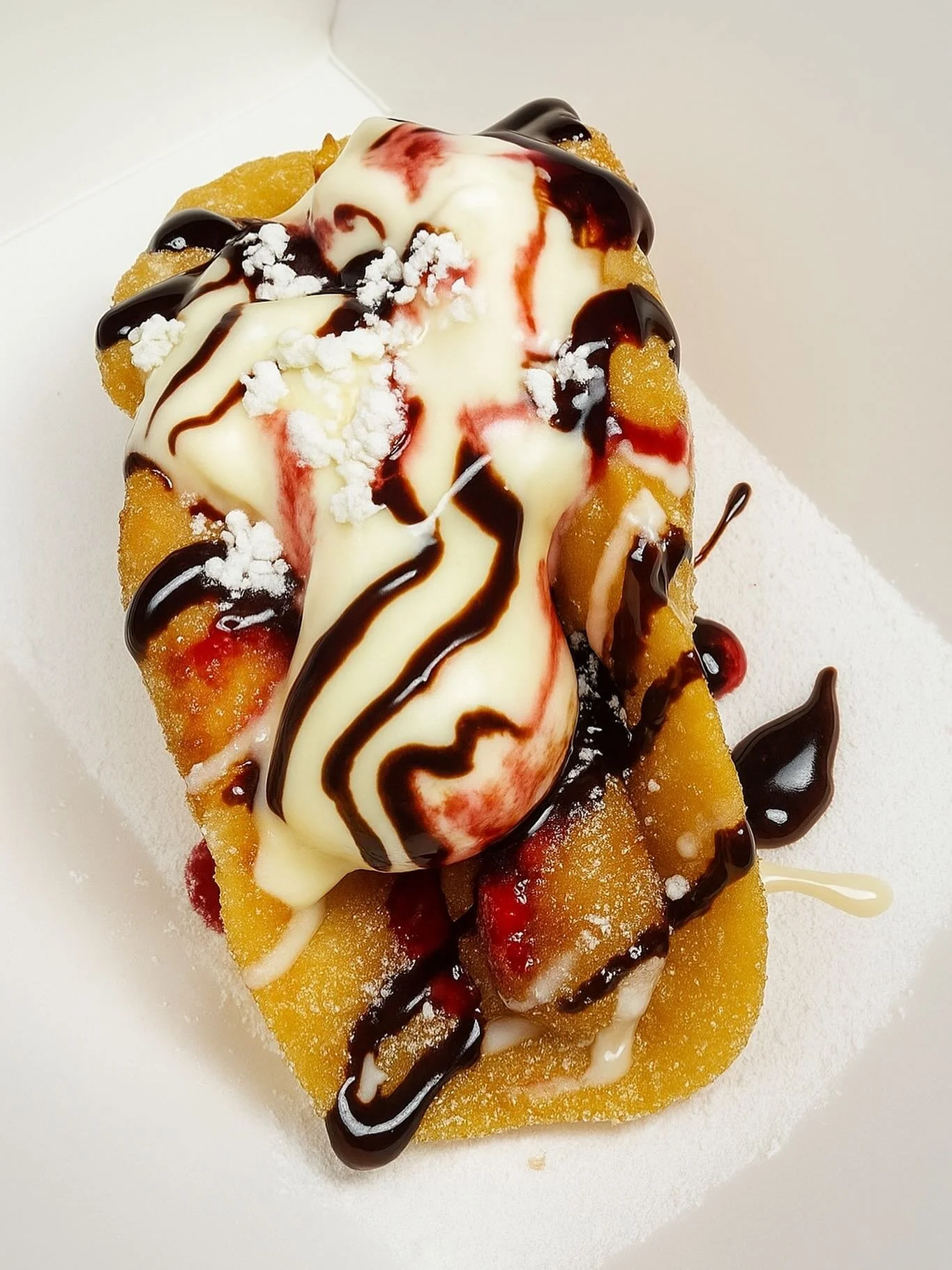 A dessert with a breaded and fried outer layer topped with vanilla and chocolate sauces, a scoop of vanilla ice cream, and garnished with powdered sugar and fruit pieces, served on a white surface.