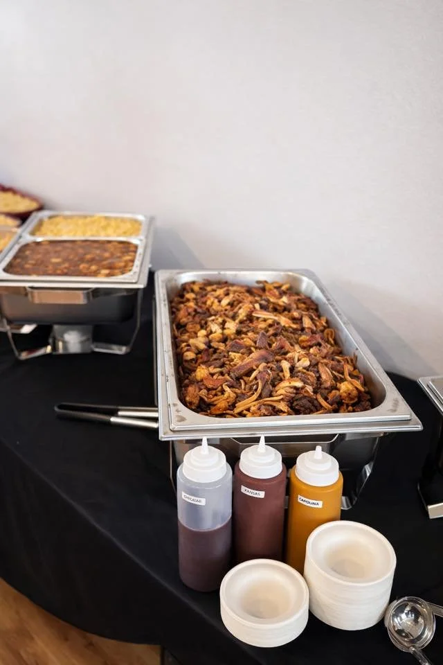 Chafing dishes containing shredded beef with onions, refried beans, and Mexican rice on a buffet table, alongside condiments and disposable bowls.