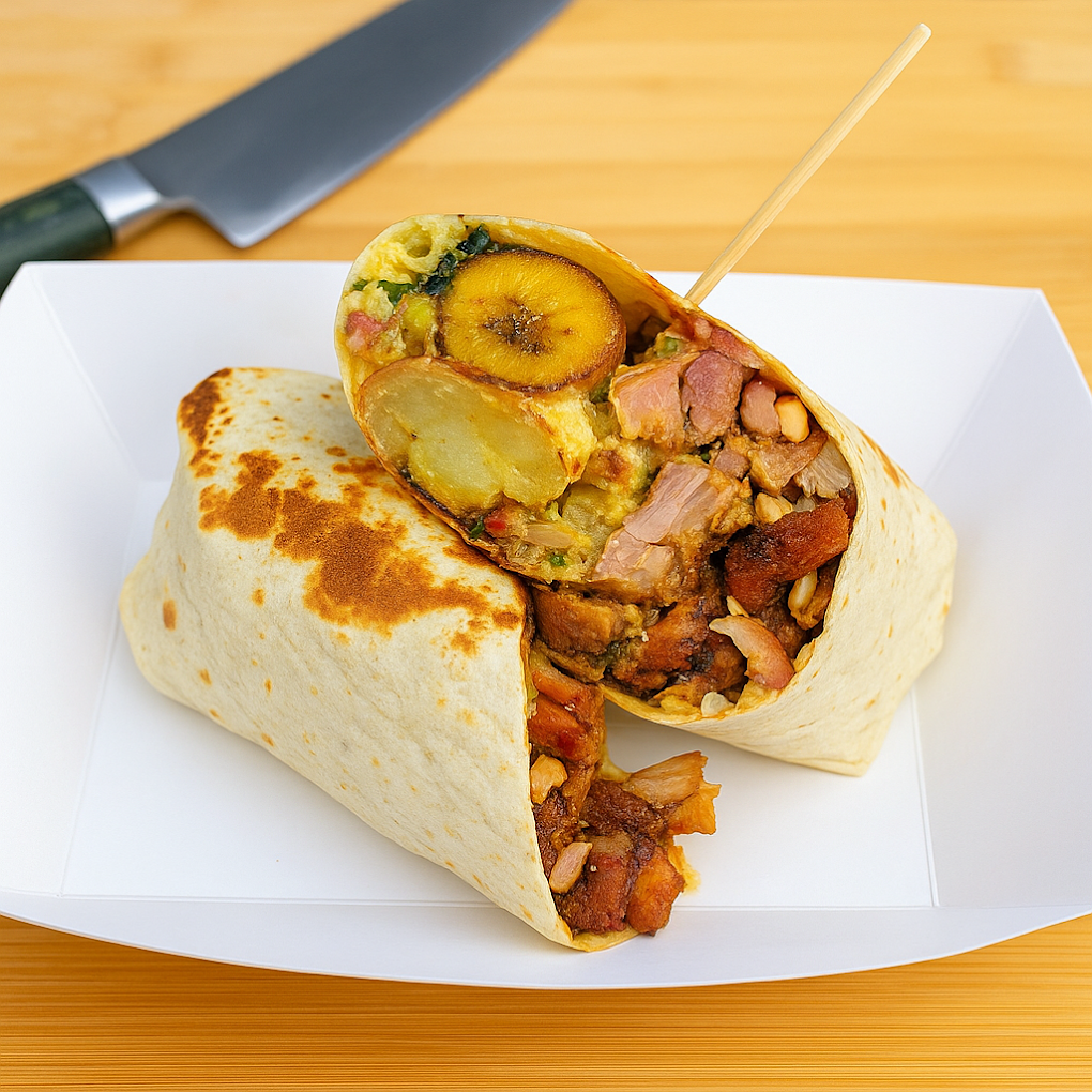 A burrito sliced in half revealing potatoes, plantains, beans, meat, and vegetables inside, served on a white tray with a knife in the background.