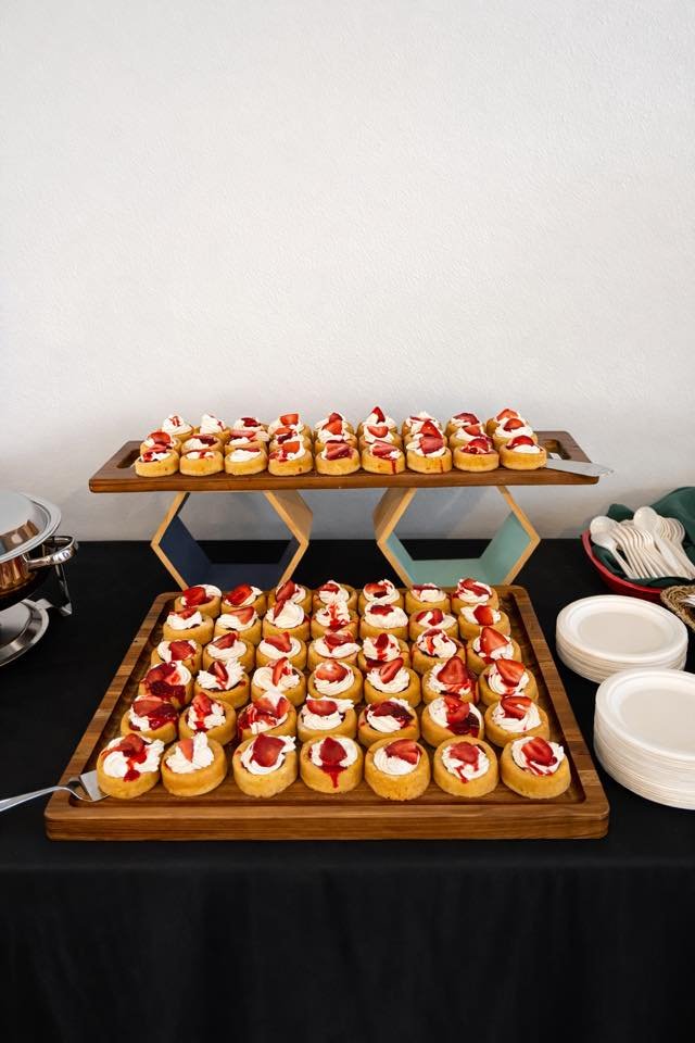 Tray of strawberry-topped mini desserts on a black table, with plates, utensils, and a white wall in the background.
