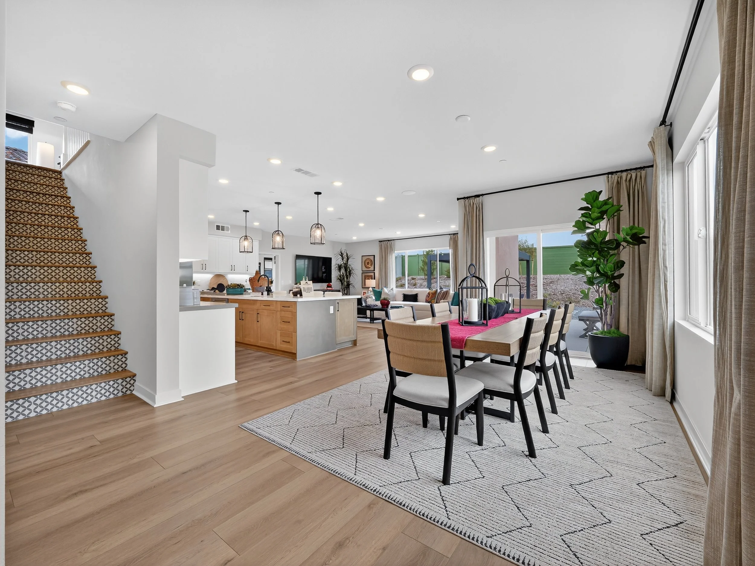 Open-concept living and dining area with hardwood floors, beige dining chairs around a table with a red table runner, large sliding glass doors with beige curtains, a tall potted plant, a kitchen island with pendant lights, and staircase with pattern