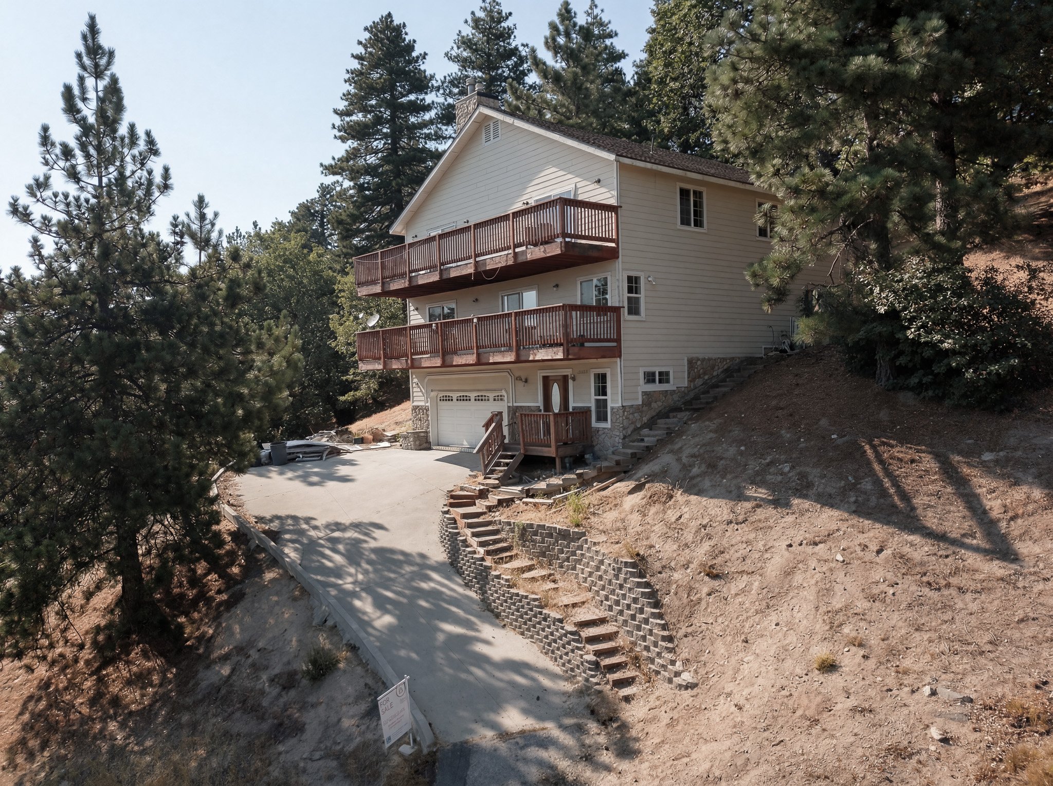 A white multi-story house with two balconies is situated on a sloped terrain surrounded by tall pine trees. There are steps and a stone retaining wall leading up to the house, with some construction materials at the base and a for sale sign nearby.
