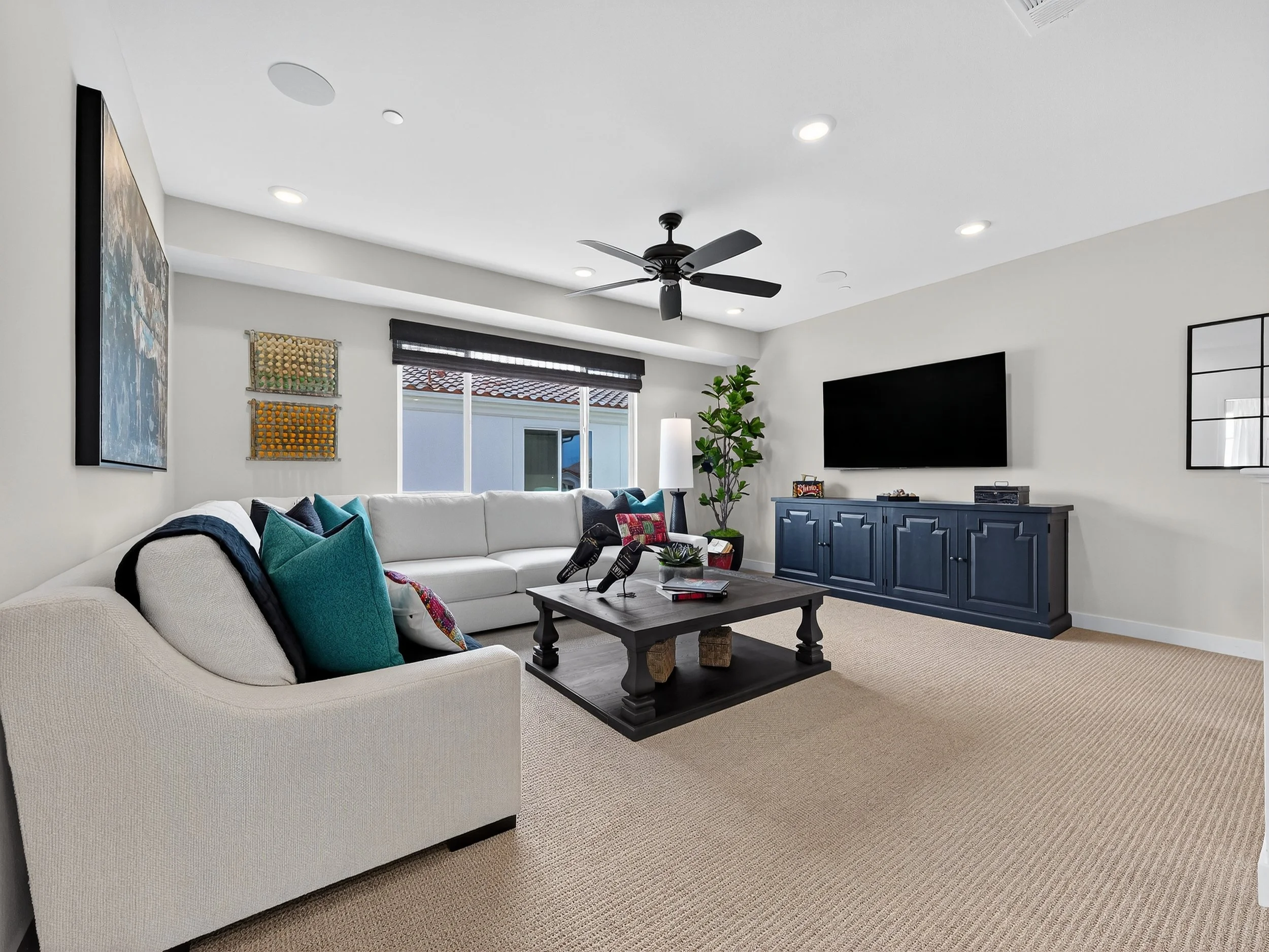 Living room with white sectional sofa with teal and patterned pillows, black coffee table, blue sideboard, wall-mounted flat-screen TV, artwork, potted plant, window with blinds, ceiling fan, and recessed lighting.