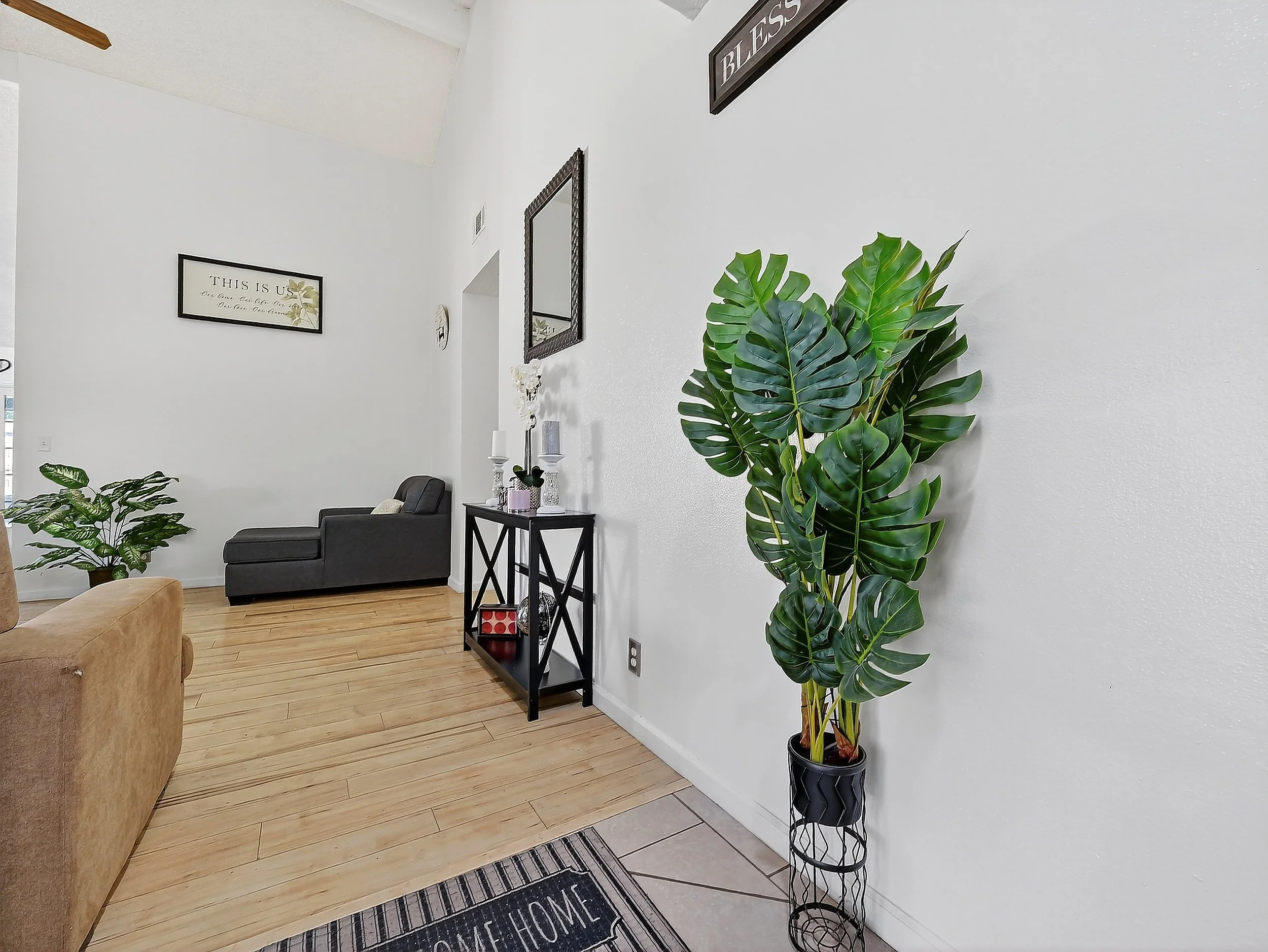 Living room with beige sofa, black chair, wooden flooring, potted plants, wall art, mirror, and a welcome mat.
