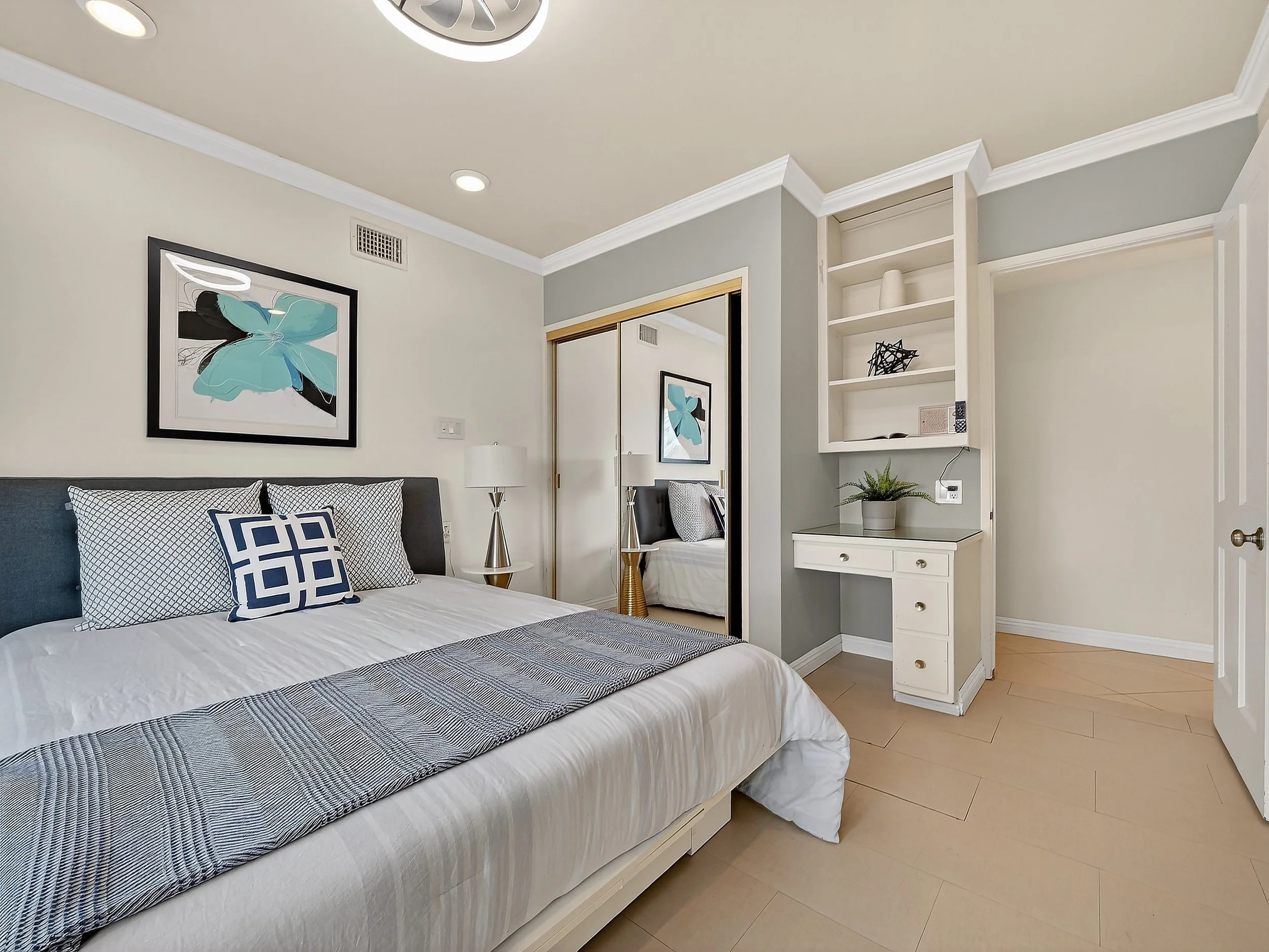 A modern bedroom with a neatly made bed, decorative pillows, framed artwork with a butterfly, a nightstand with a lamp, mirrored closet doors, built-in white shelves with decorative objects and a small plant, light-colored hardwood floors, and neutra