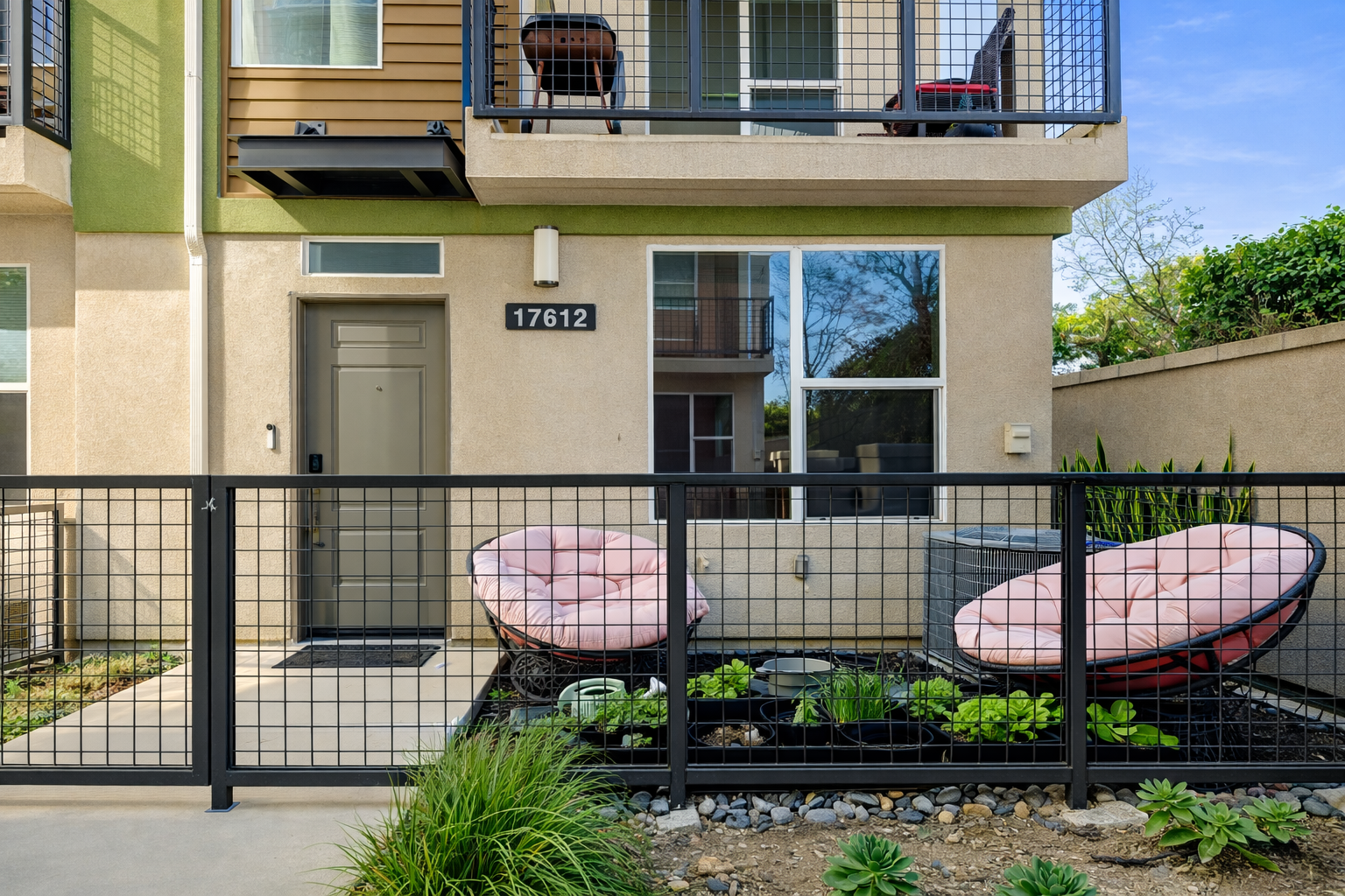 Apartment patio with pink cushioned chairs, plants, and black metal fence, with the apartment door and window overhead.