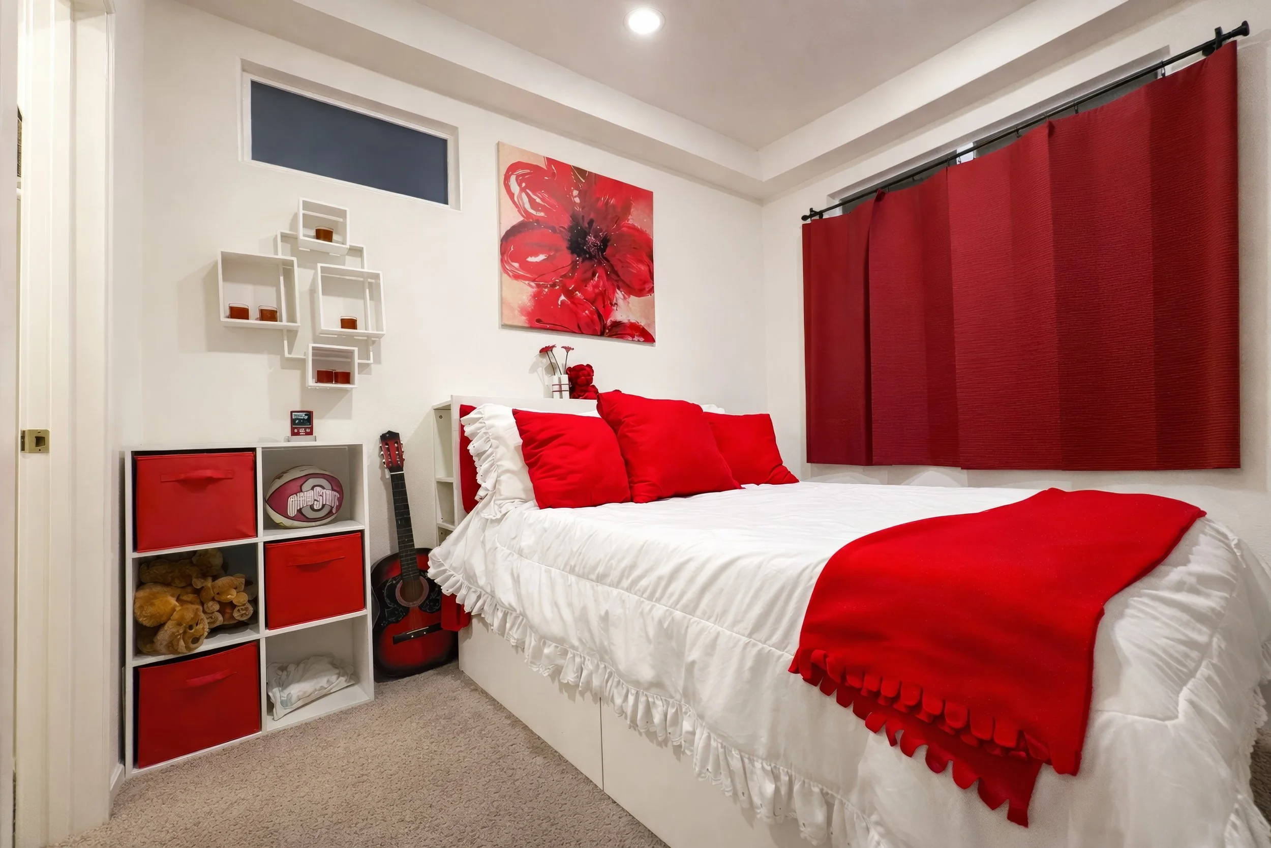A bedroom decorated in red and white, featuring a bed with white ruffled bedding and multiple red pillows. Above the bed is a red floral painting. To the right, there is a window covered by red curtains. To the left, white shelves hold small candles 