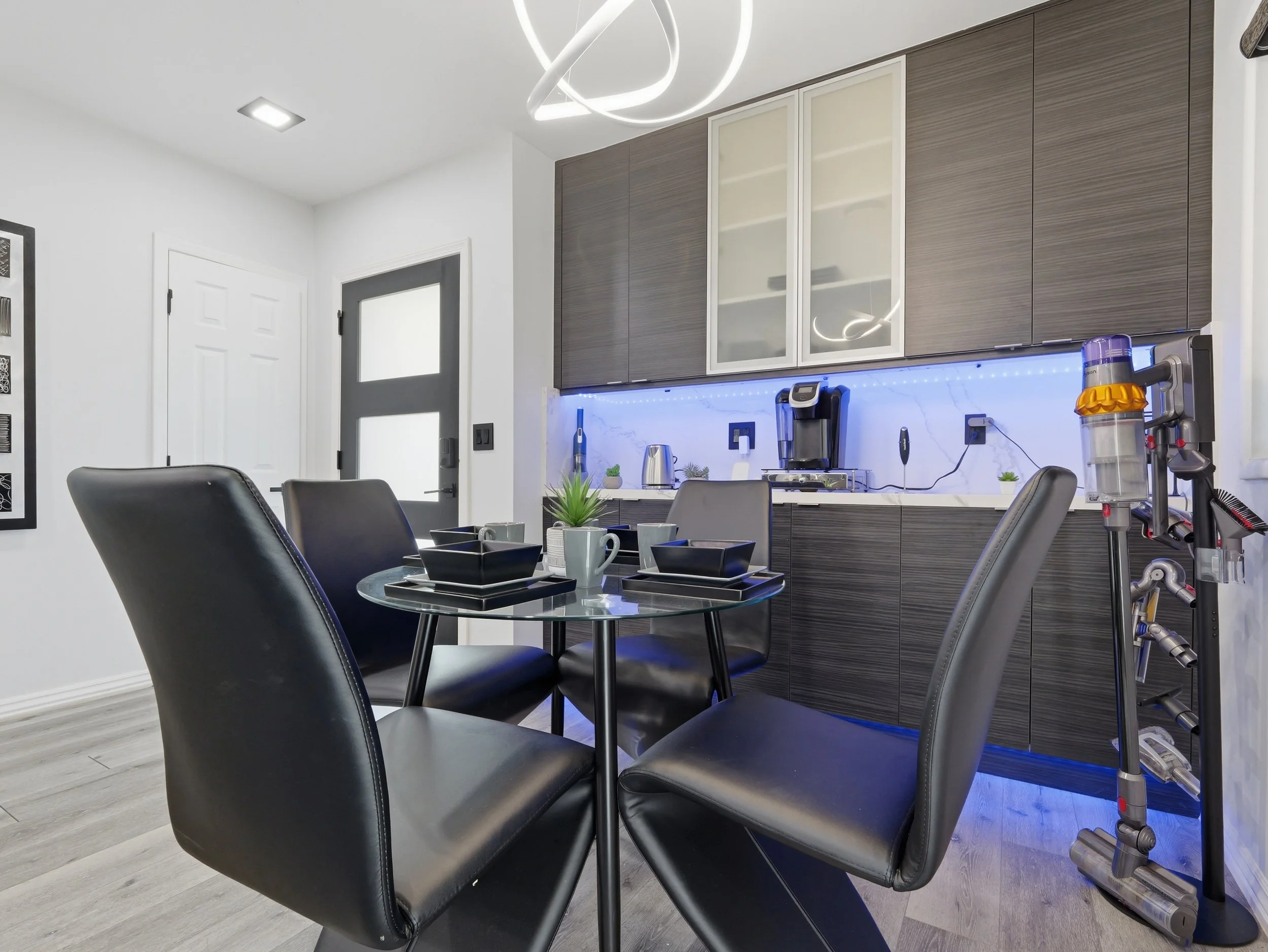 A modern kitchen with black chairs around a round glass table, white cabinetry with integrated lighting, and a wall with a water filter and coffee maker. There are plants and cups on the table, and a contemporary light fixture on the ceiling.