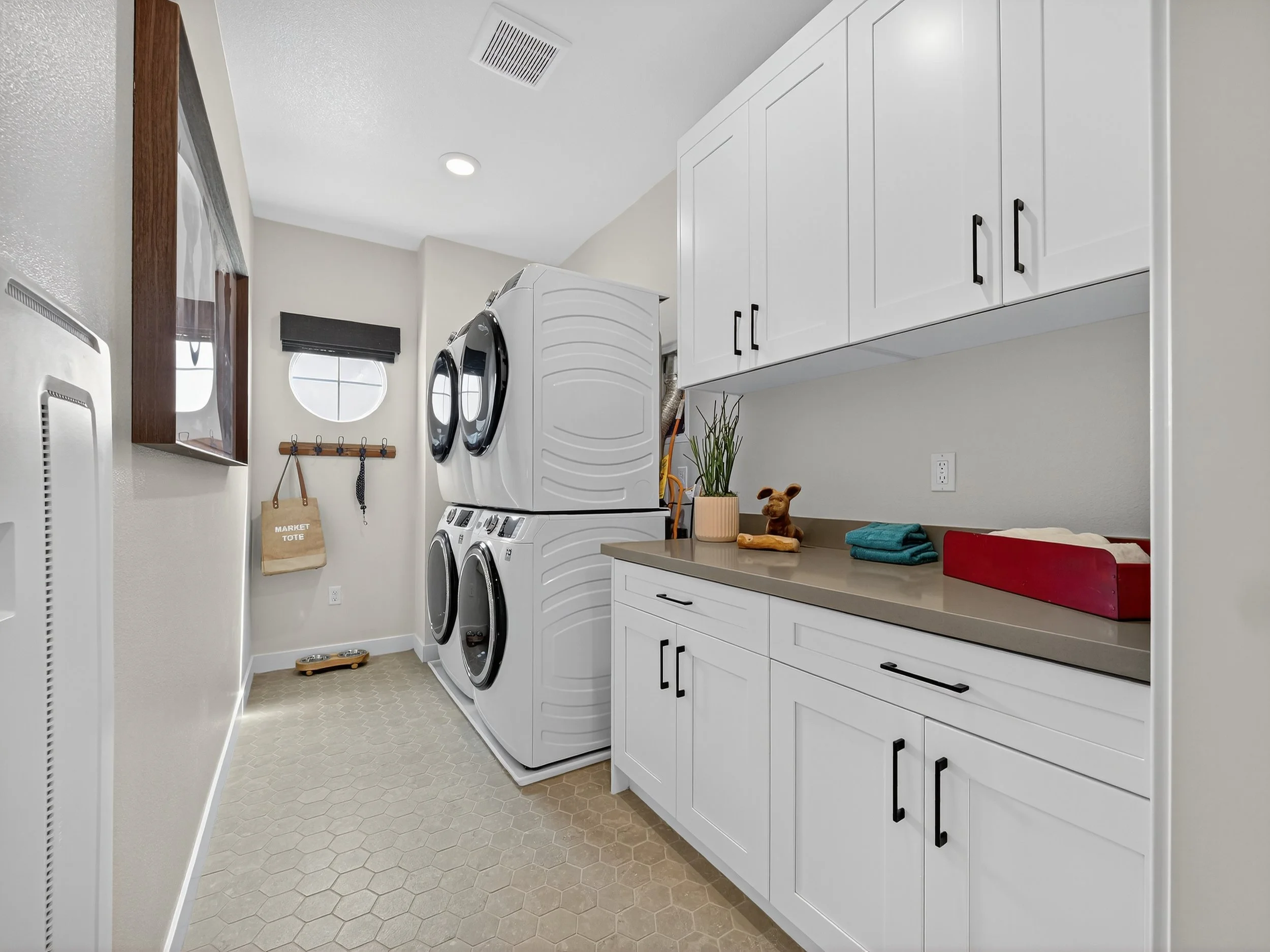 A laundry room with a front-loading washer and dryer stacked, white cabinets above, a countertop with folded towels and a decorative plant, a wall hook with bags, and a small window with a black shade.