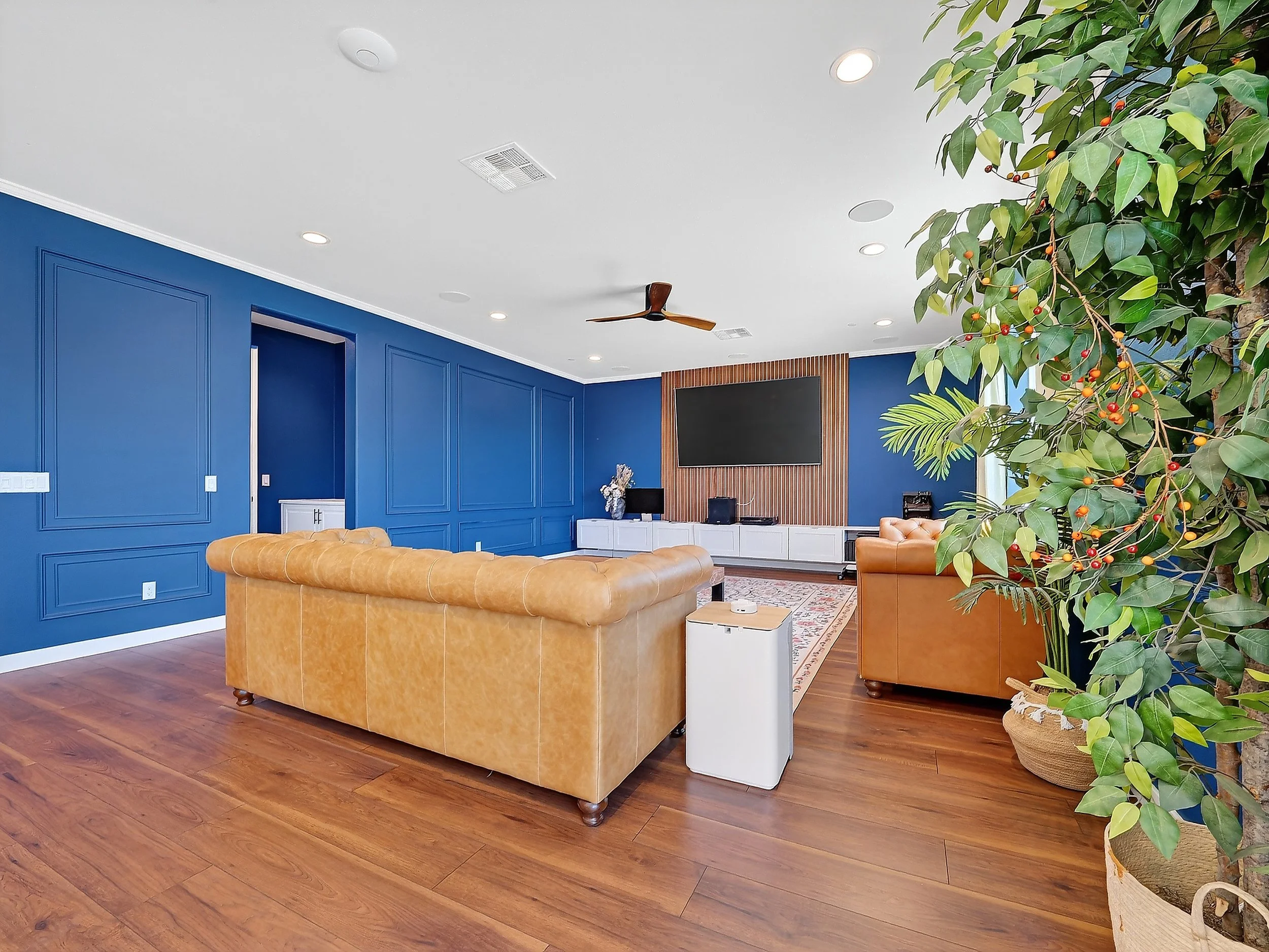 Living room with blue accent wall, tan leather sofas, a large flat-screen TV on a wooden panel, white entertainment console, ceiling fan, wooden floors, and a large potted plant.