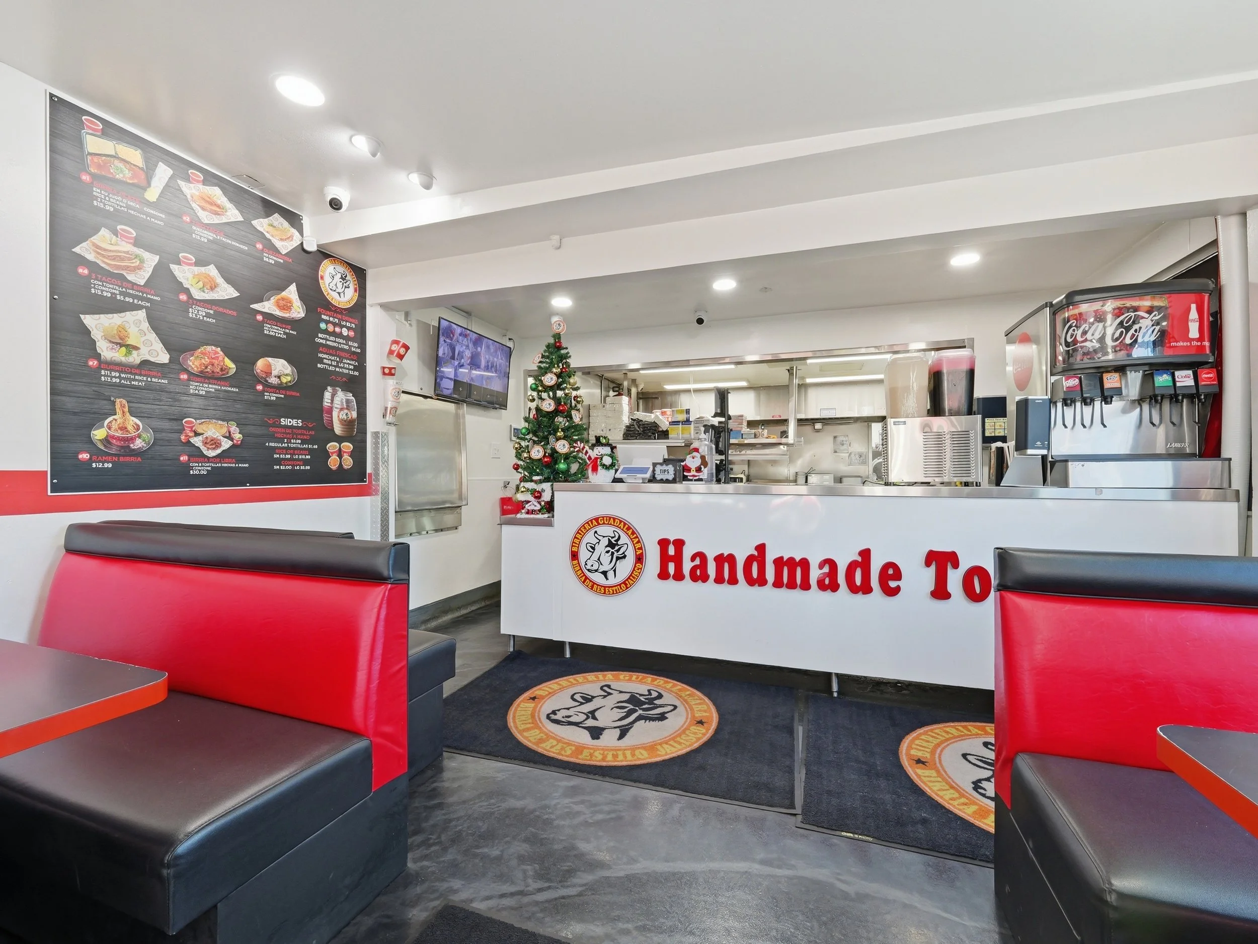 Inside a fast-food restaurant decorated for Christmas, with red and black seating, a Christmas tree, and a large menu board on the wall showing various food items.