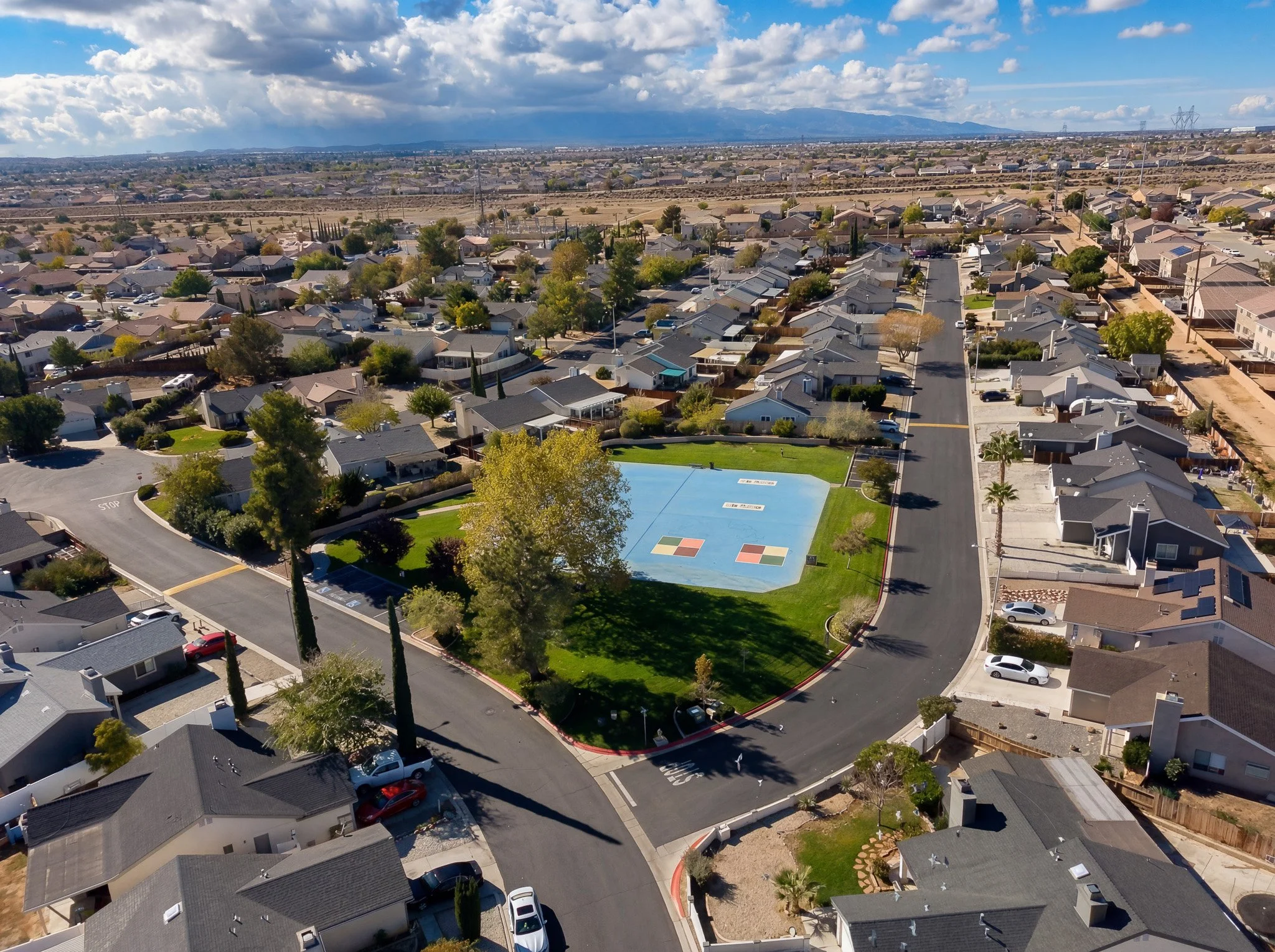 Aerial view of a suburban neighborhood with houses, trees, and streets, with mountains in the background under a partly cloudy sky.