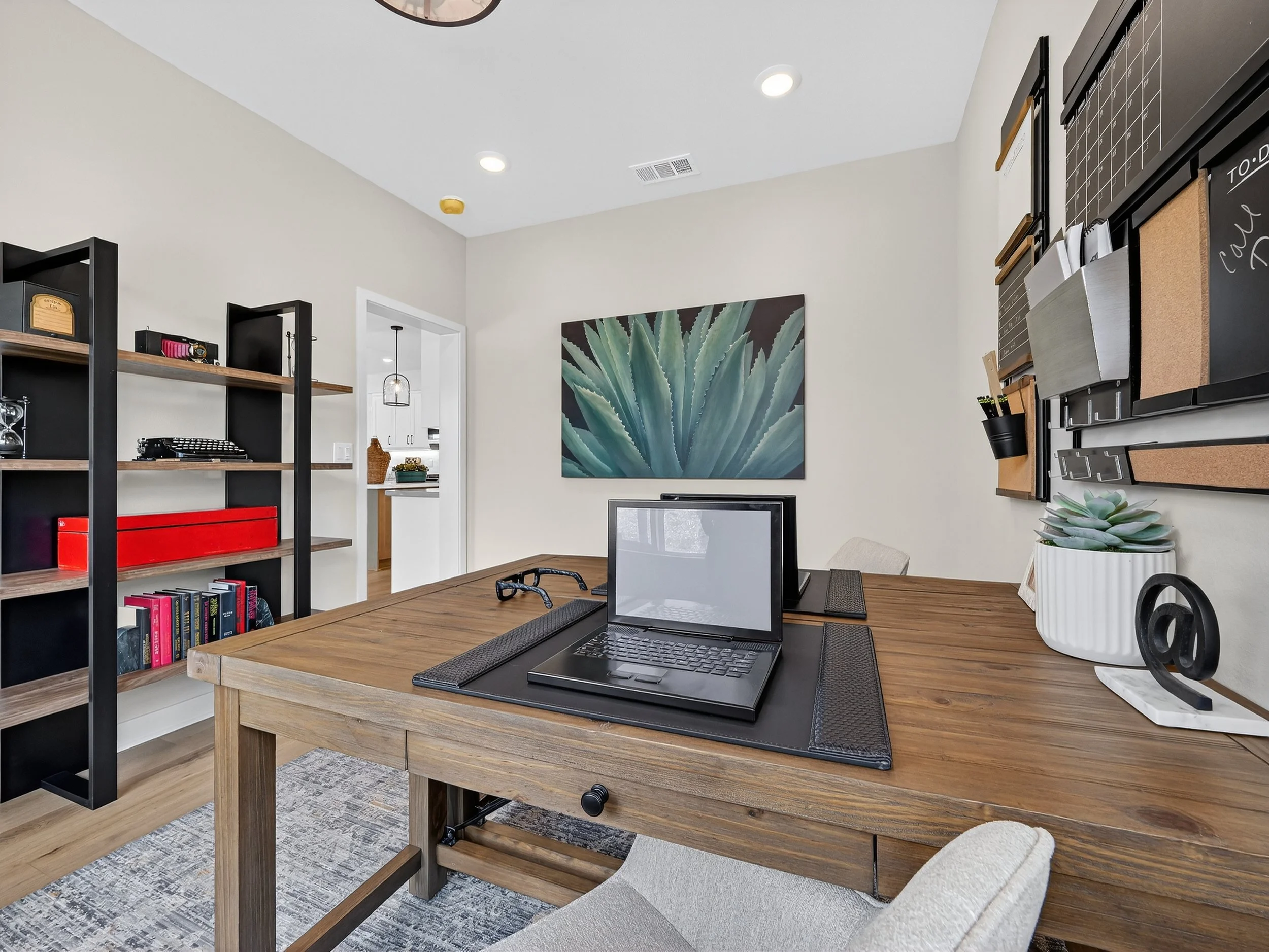 A modern home office with a wooden desk, a laptop, a potted succulent plant, a black mesh mouse pad, and decorative shelving, with a large cactus-themed painting on the wall.