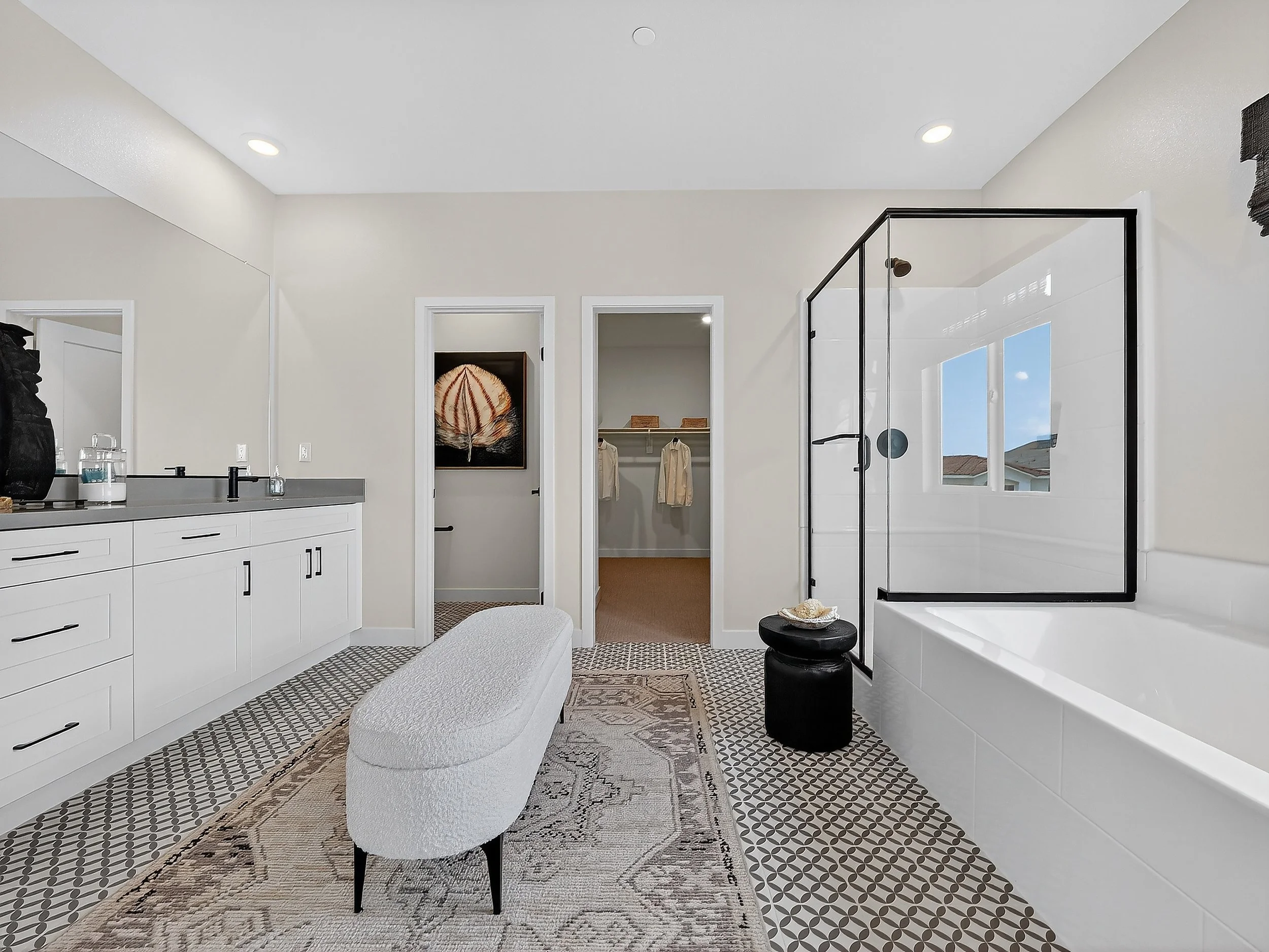 Modern bathroom with white cabinets, a patterned rug, a white rounded chair, a black side table, a glass shower enclosure, a window, and a walk-in closet with hanging clothes.