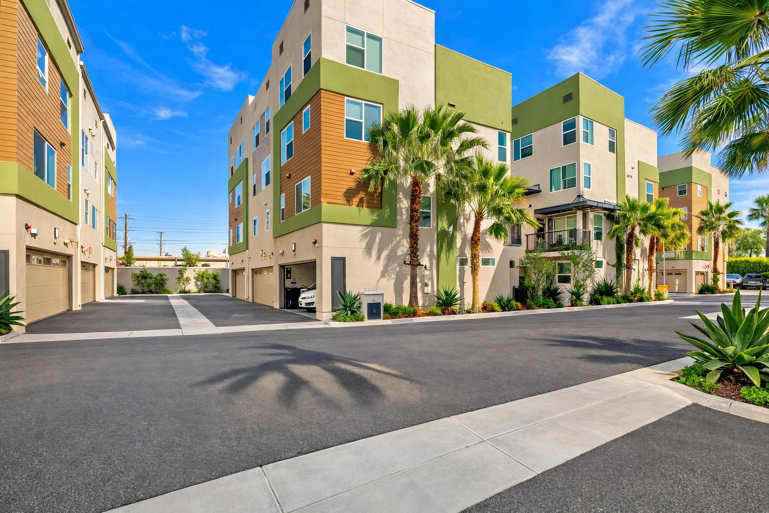 Colorful modern apartment complex with green, beige, and wood-paneled sections, surrounded by palm trees and desert plants, under a bright blue sky with some clouds.