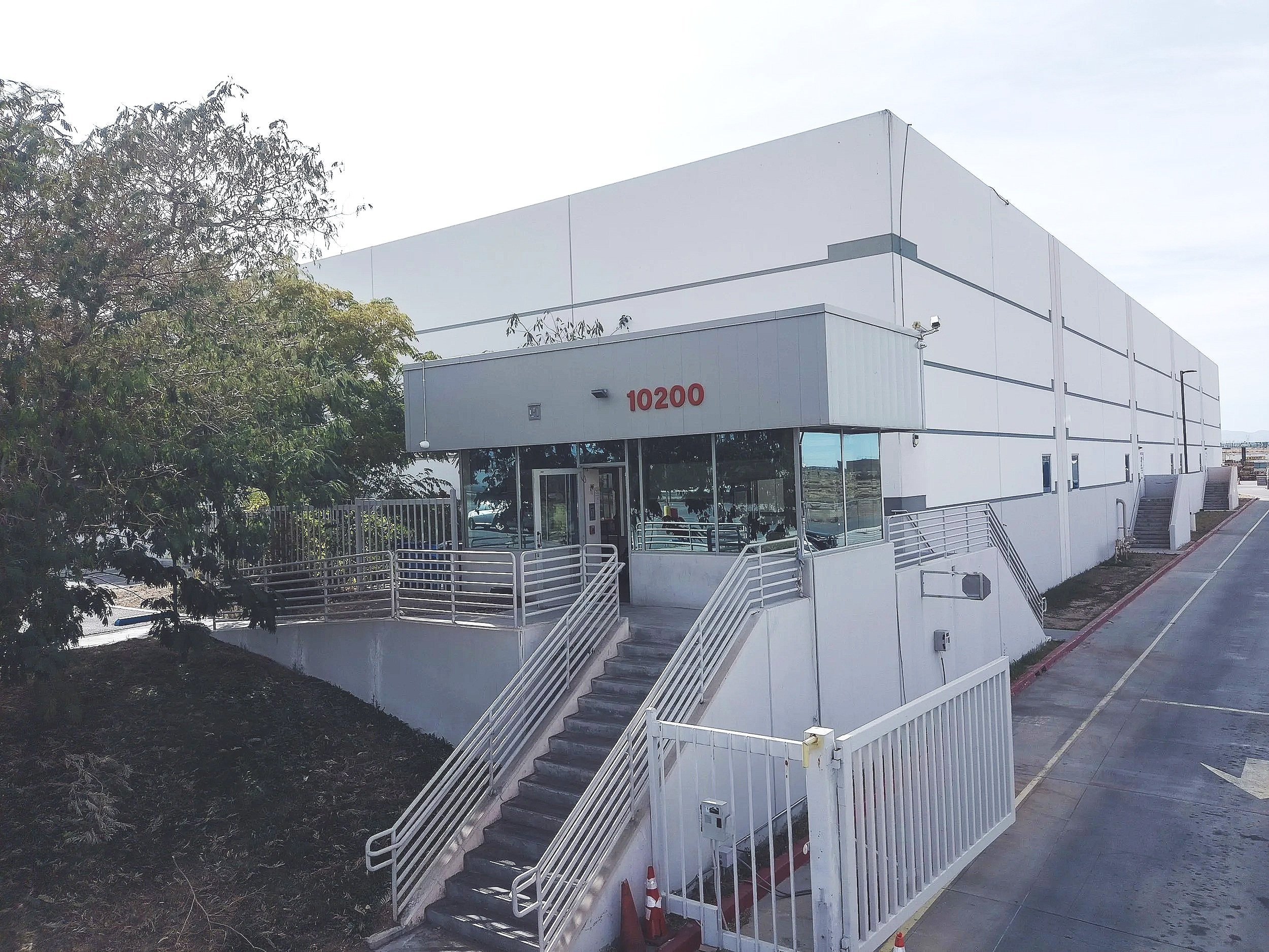 A white multi-story building with the number 10200 in red on the side, featuring an exterior staircase with railings leading up to a door, surrounded by trees and a parking lot.