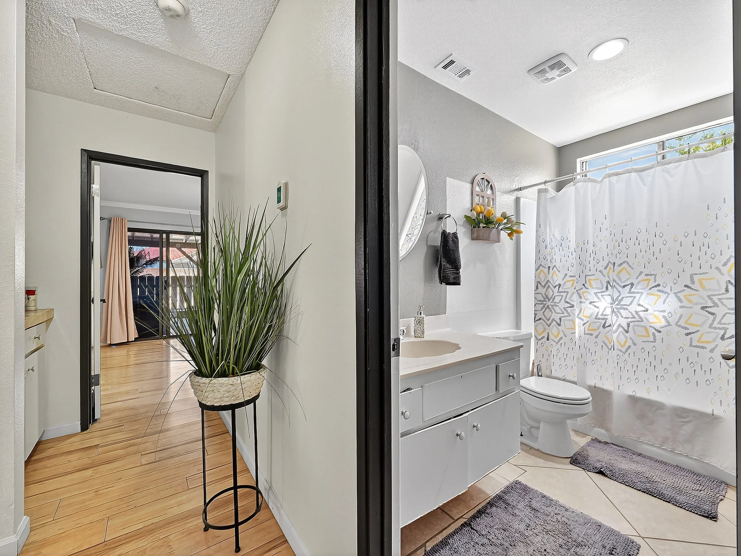 Split view of a home interior: left side shows a hallway with wooden flooring, a potted plant, and a doorway leading to a room with pink curtains and sliding glass doors; right side shows a bathroom with a vanity, mirror, toilet, and a shower with a 