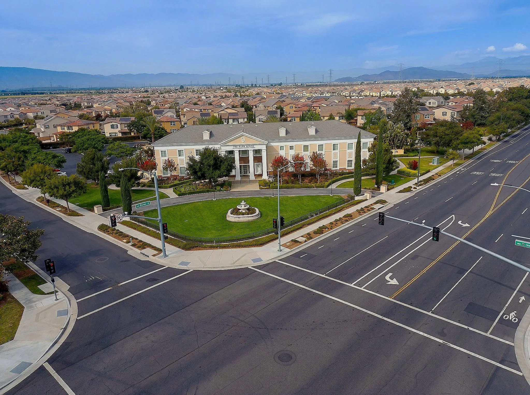 Aerial view of a suburban neighborhood with uniform houses, a large white building with columns labeled "The Park House," trees, and a landscaped circular fountain at an intersection with traffic lights. In the background, mountains are visible under a cloudy sky.