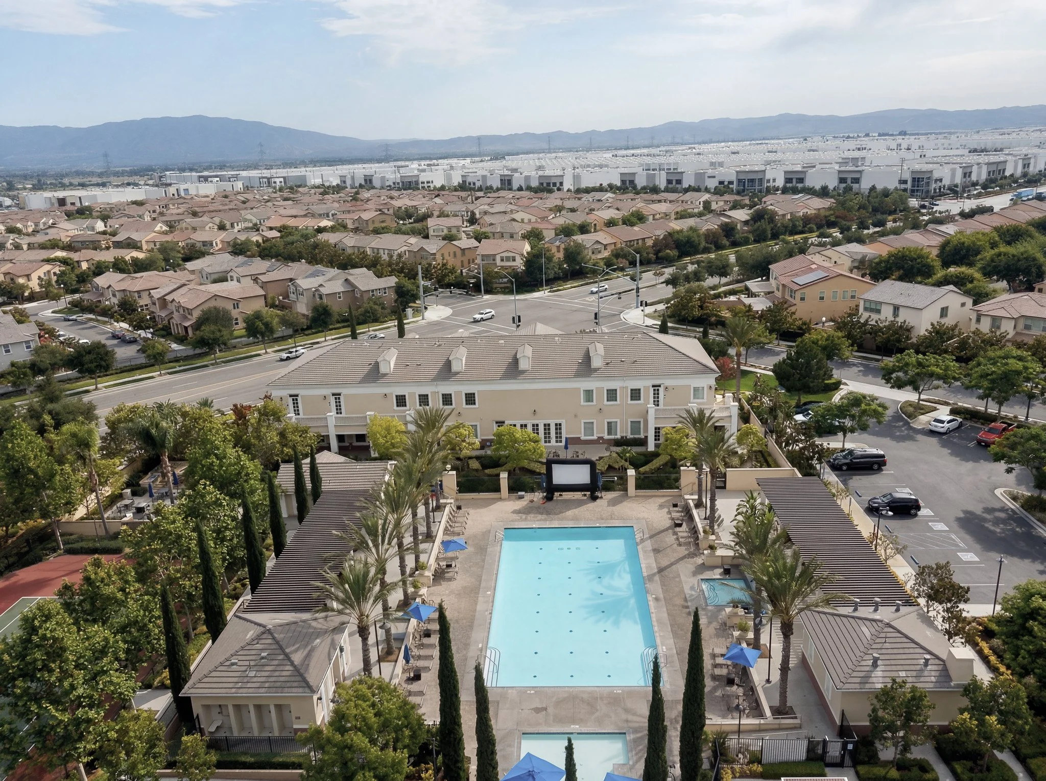 Aerial view of a residential area with houses, trees, and two swimming pools in the foreground, with a large industrial or warehouse complex and mountains in the background.