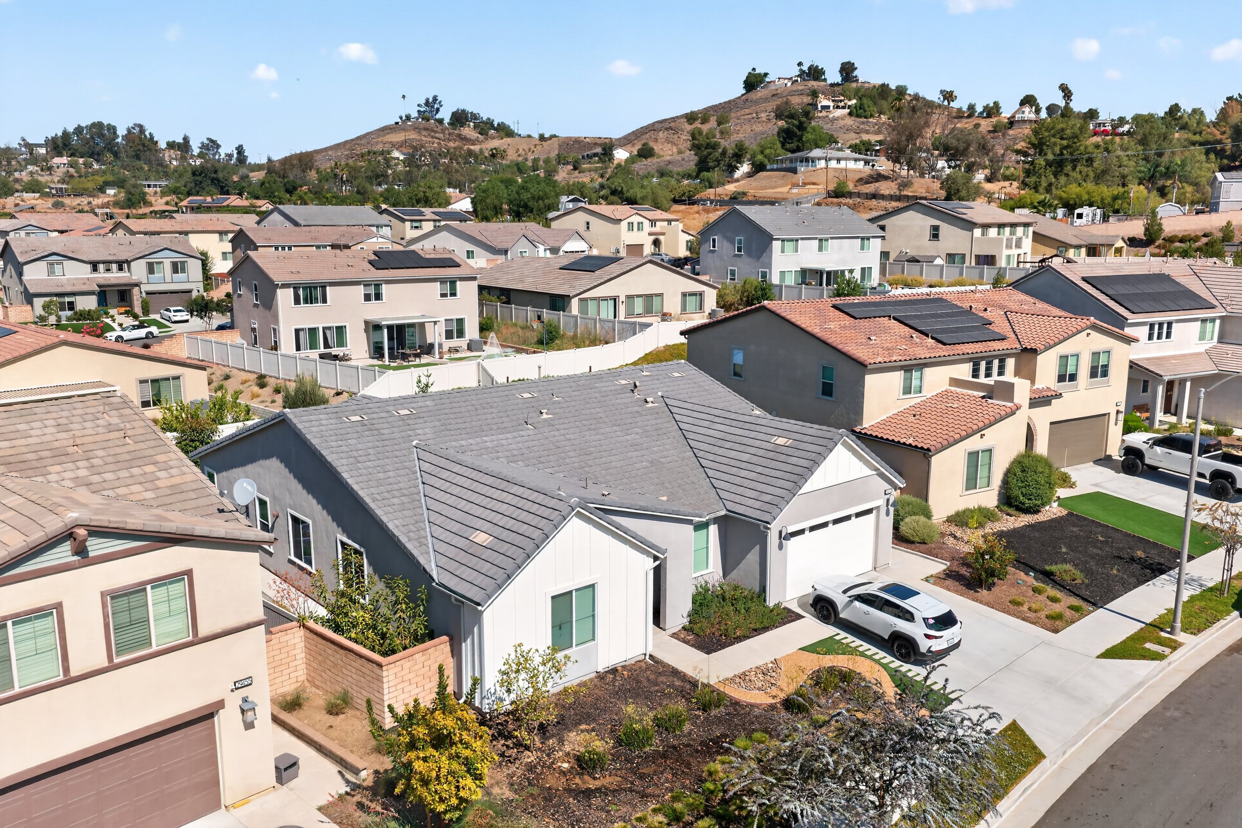 A residential neighborhood with multiple two-story houses featuring tile and shingle roofs, some with solar panels, in a hilly area with a backdrop of dry, grassy hills and some trees under a blue sky.