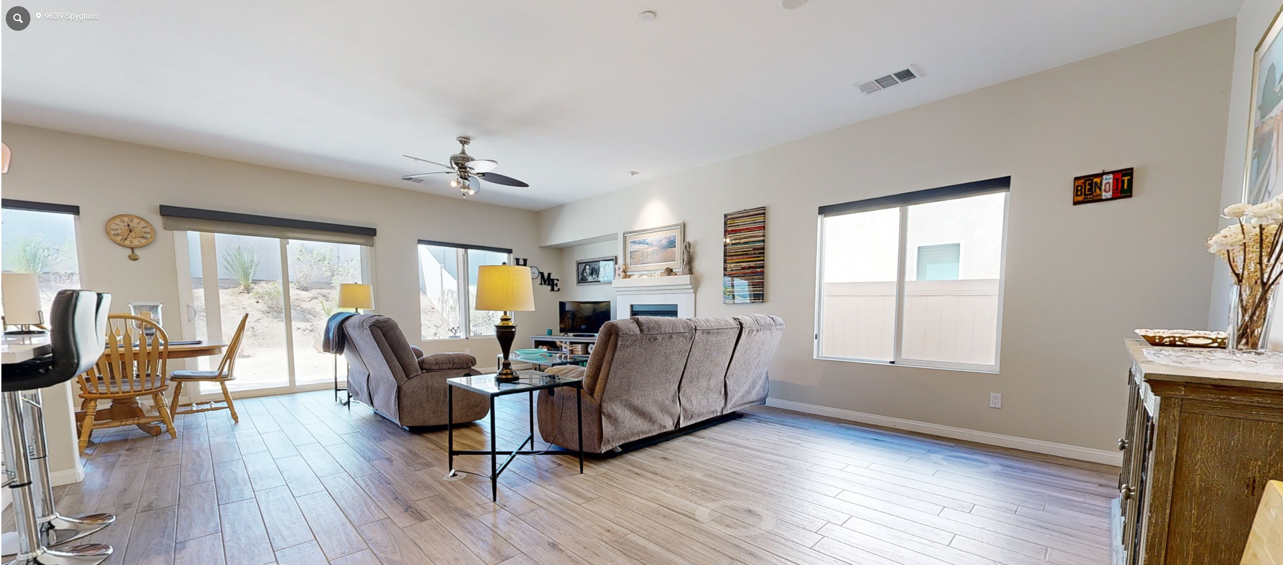 Living room with beige sectional sofa, wooden dining table with chairs, windows with view of backyard, ceiling fan, and various decorative items on walls and mantle.