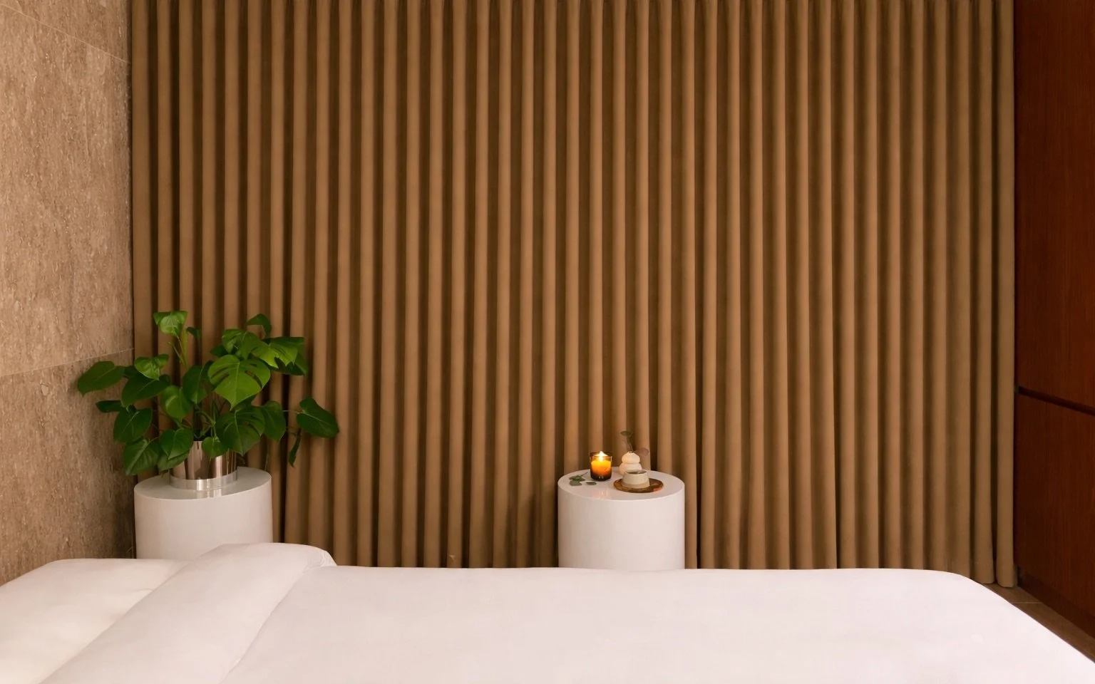 A bedroom with a bed, a green plant in a white pot, a small white cylindrical table with a candle and decor, and a brown curtain.