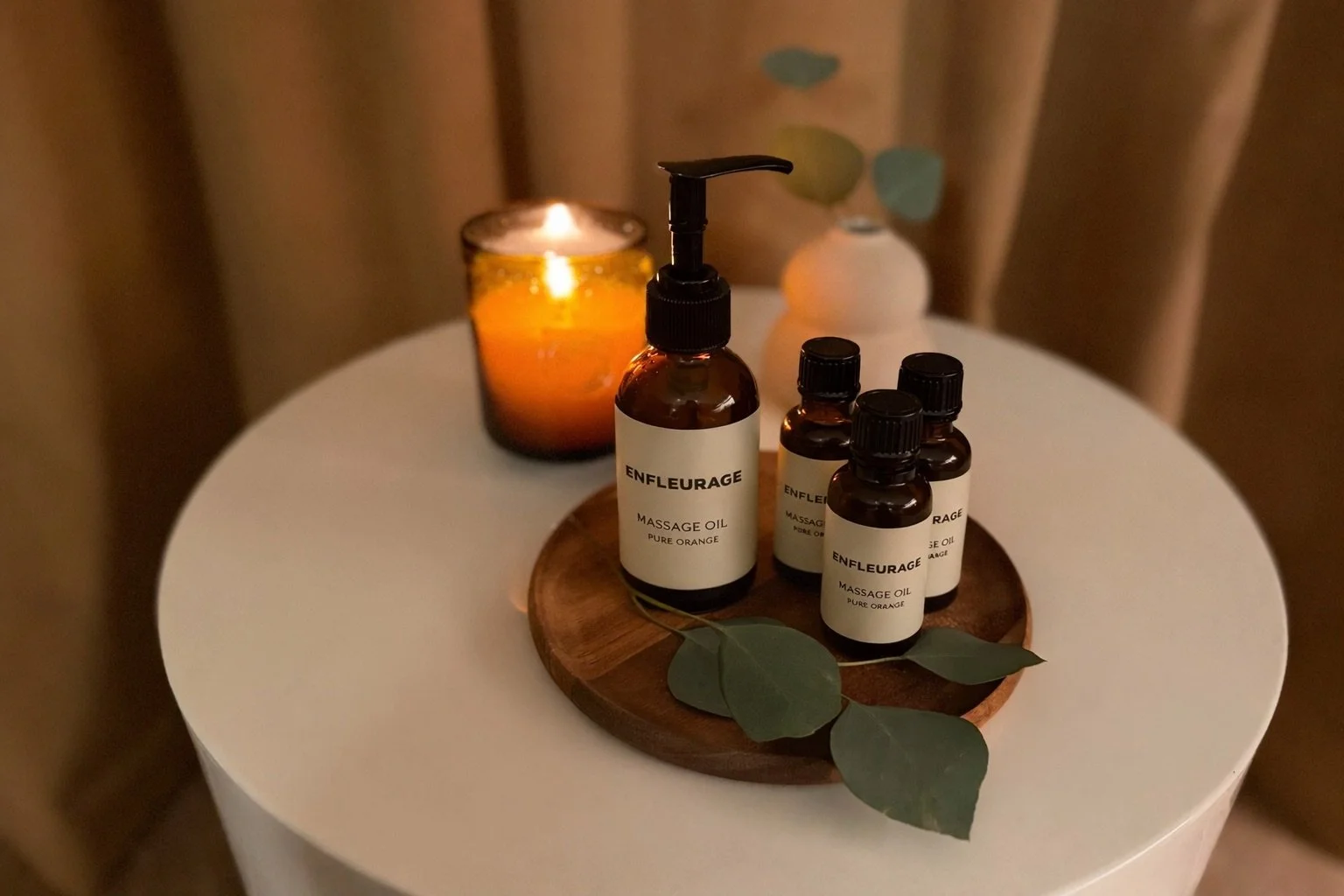 A round white table with a wooden tray holding four brown bottles labeled 'ENFLEURAGE Massage Oil Pure Orange' and a few eucalyptus leaves. A lit candle and a small white vase with eucalyptus branches are in the background.