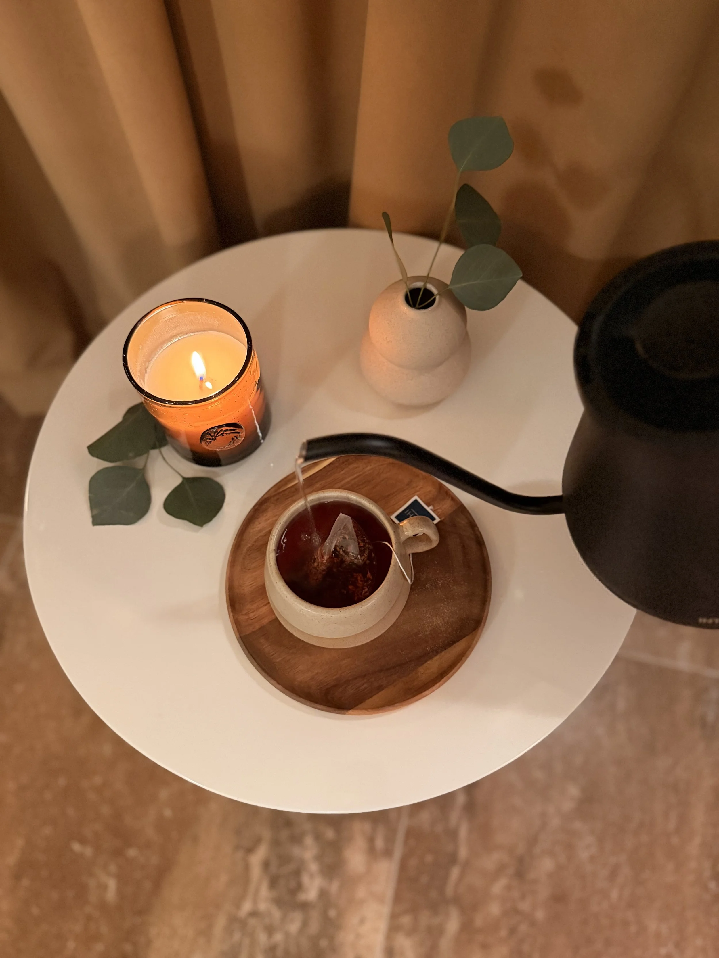 A white table with a black teapot pouring hot water into a mug of tea with a tea bag, a lit candle in an orange holder, and a beige vase with green eucalyptus leaves.