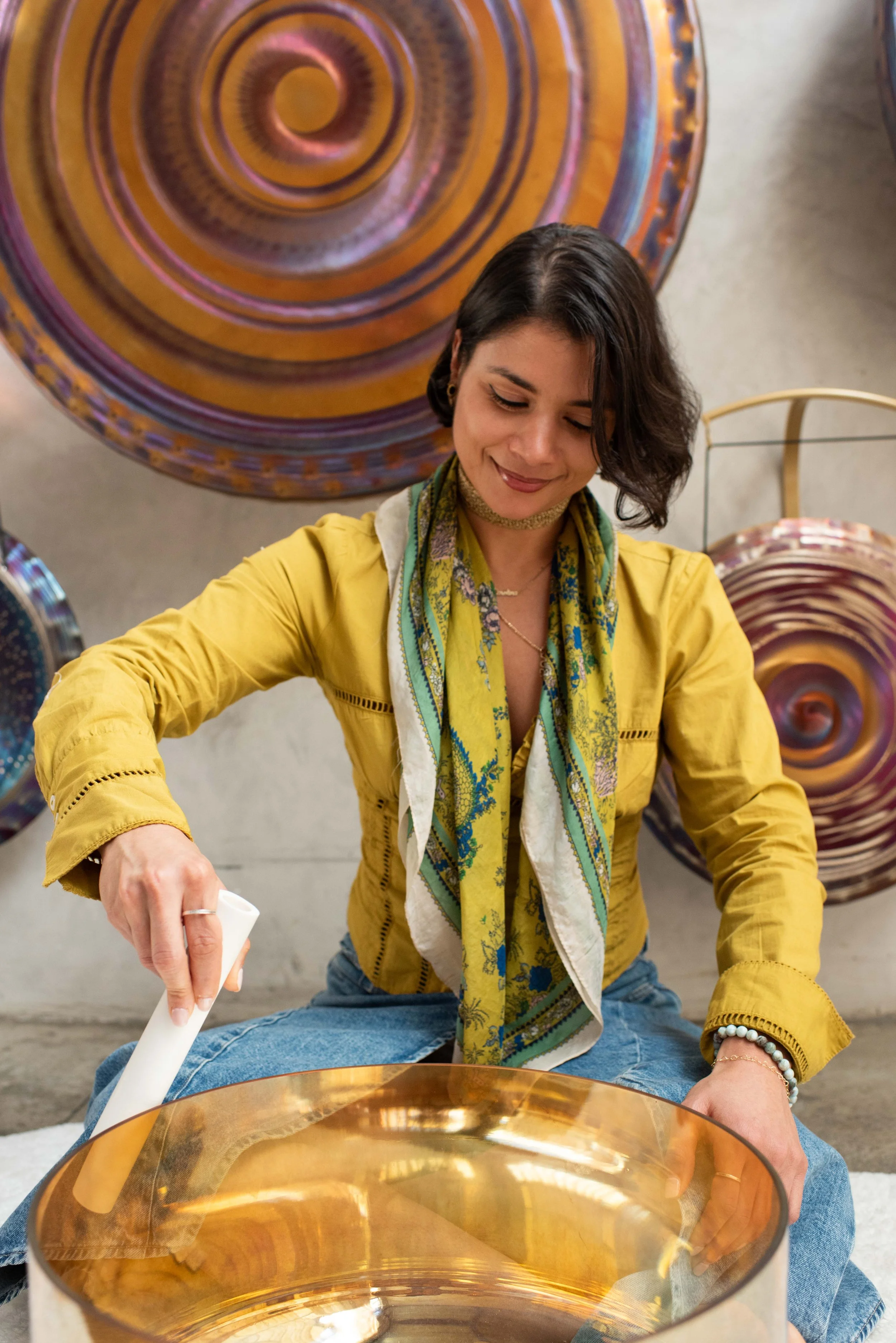 A woman with dark hair, wearing a yellow jacket and a colored scarf, is sitting on the floor and shaping a large, translucent amber-colored glass art piece.