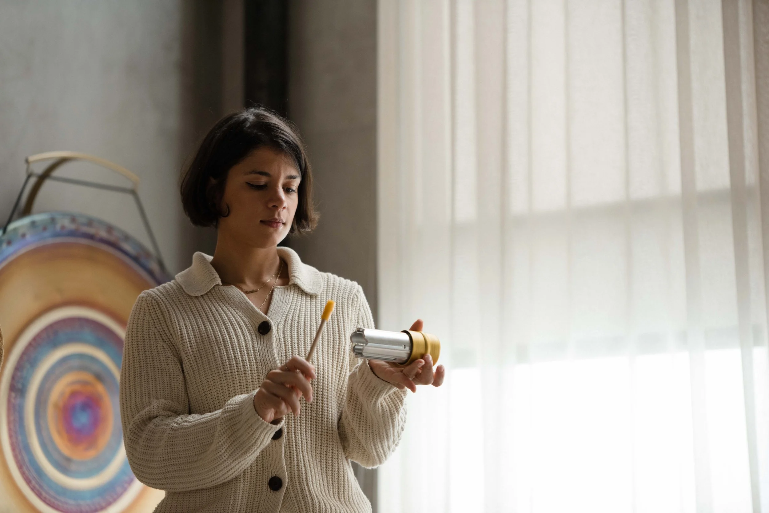 A woman holding a metallic object and a small yellow-handled tool, standing near a colorful circular artwork on the wall and a window with light curtains.