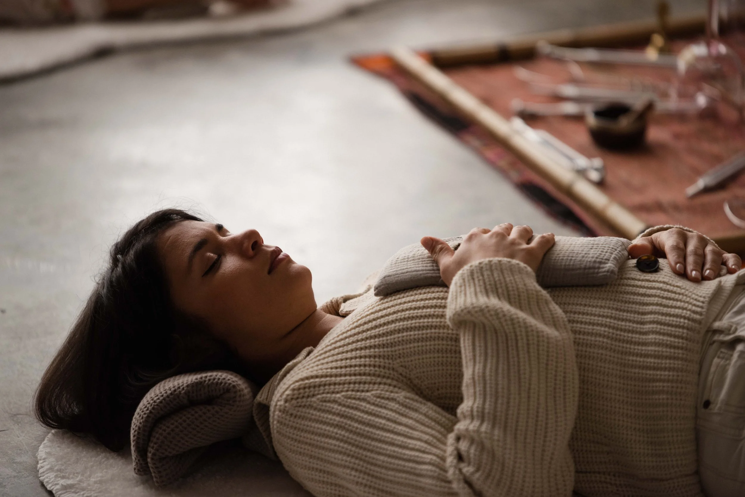 A woman lying on her back on a concrete floor with eyes closed, resting her head on a rolled-up towel, wearing a beige sweater, with a toolbox and tools in the background.