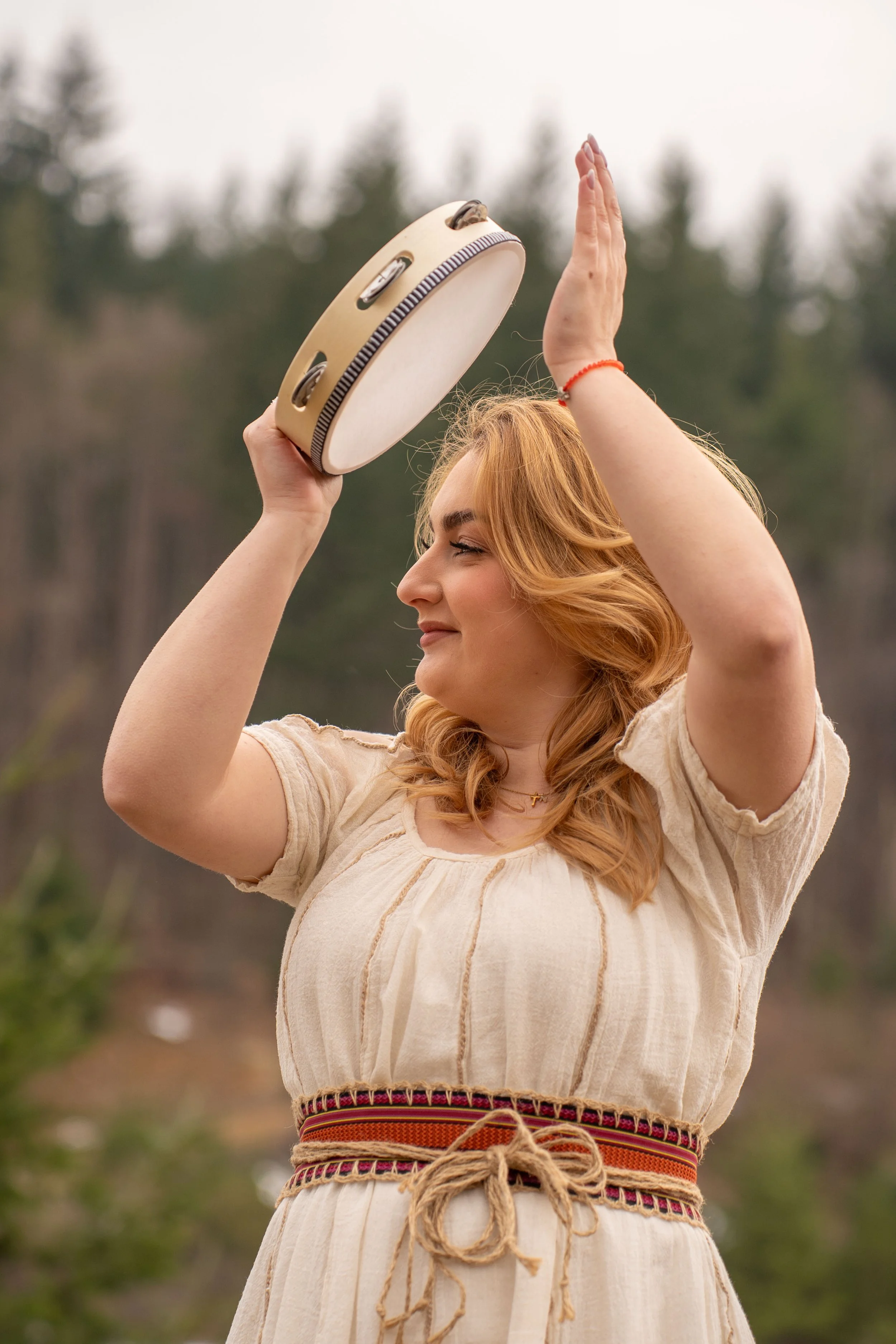 Woman with curly blonde hair standing outdoors, holding a tambourine in the air, smiling, wearing a beige dress and a woven belt, with trees and cloudy sky in the background.