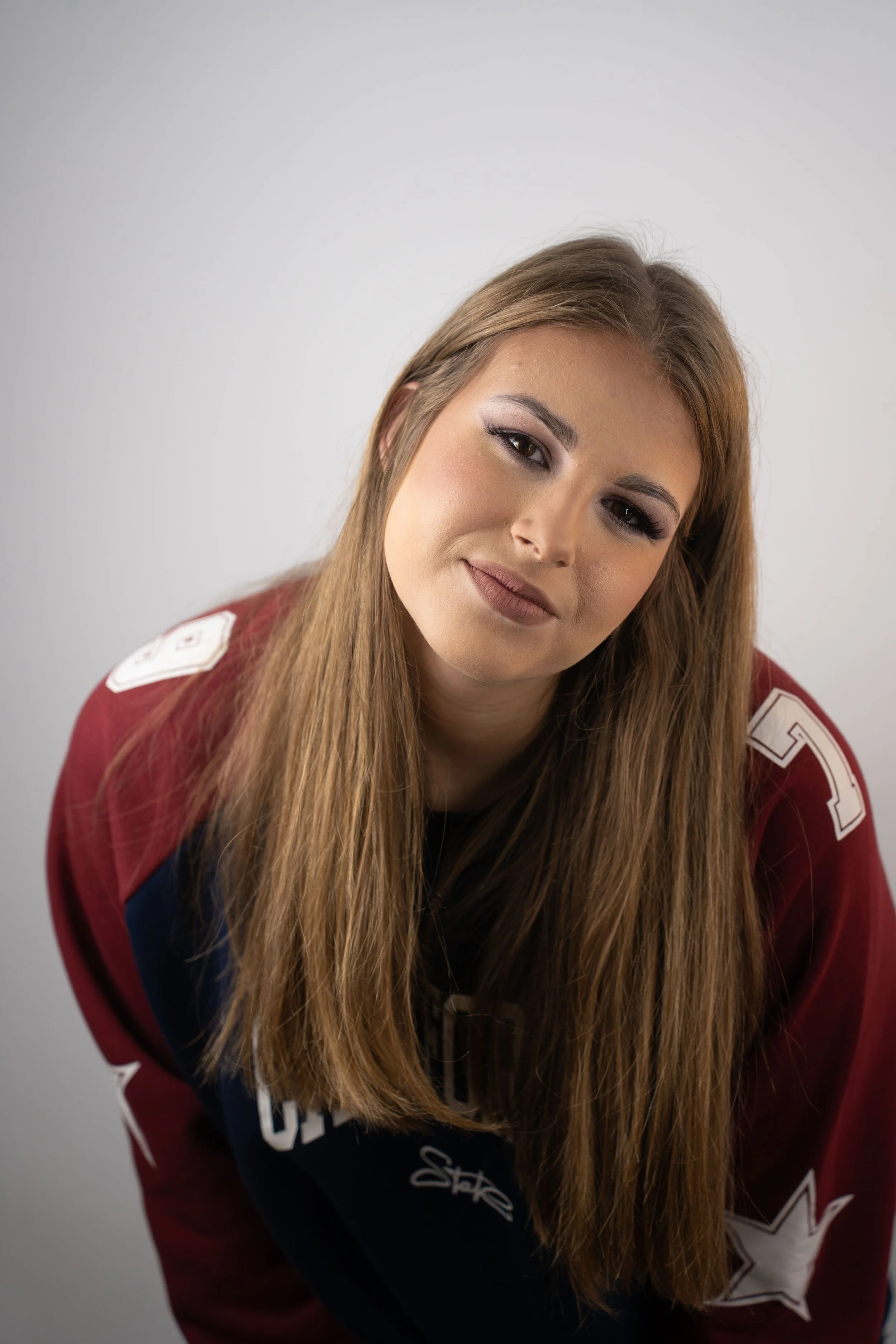 Close-up of a young woman with long, straight light brown hair, wearing a red, white, and blue sports jersey, posing against a plain, light gray background.