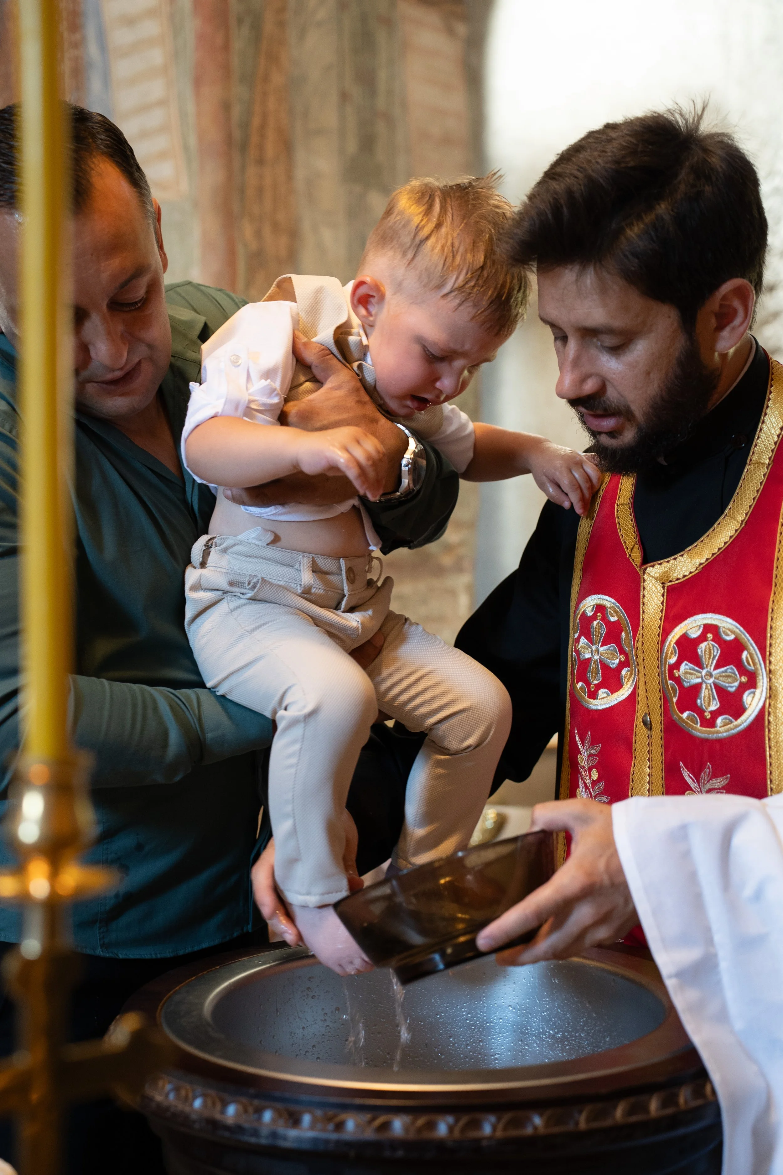 A young boy being blessed by a priest during a baptism ceremony in a church, with water being poured onto his head over a baptismal font.