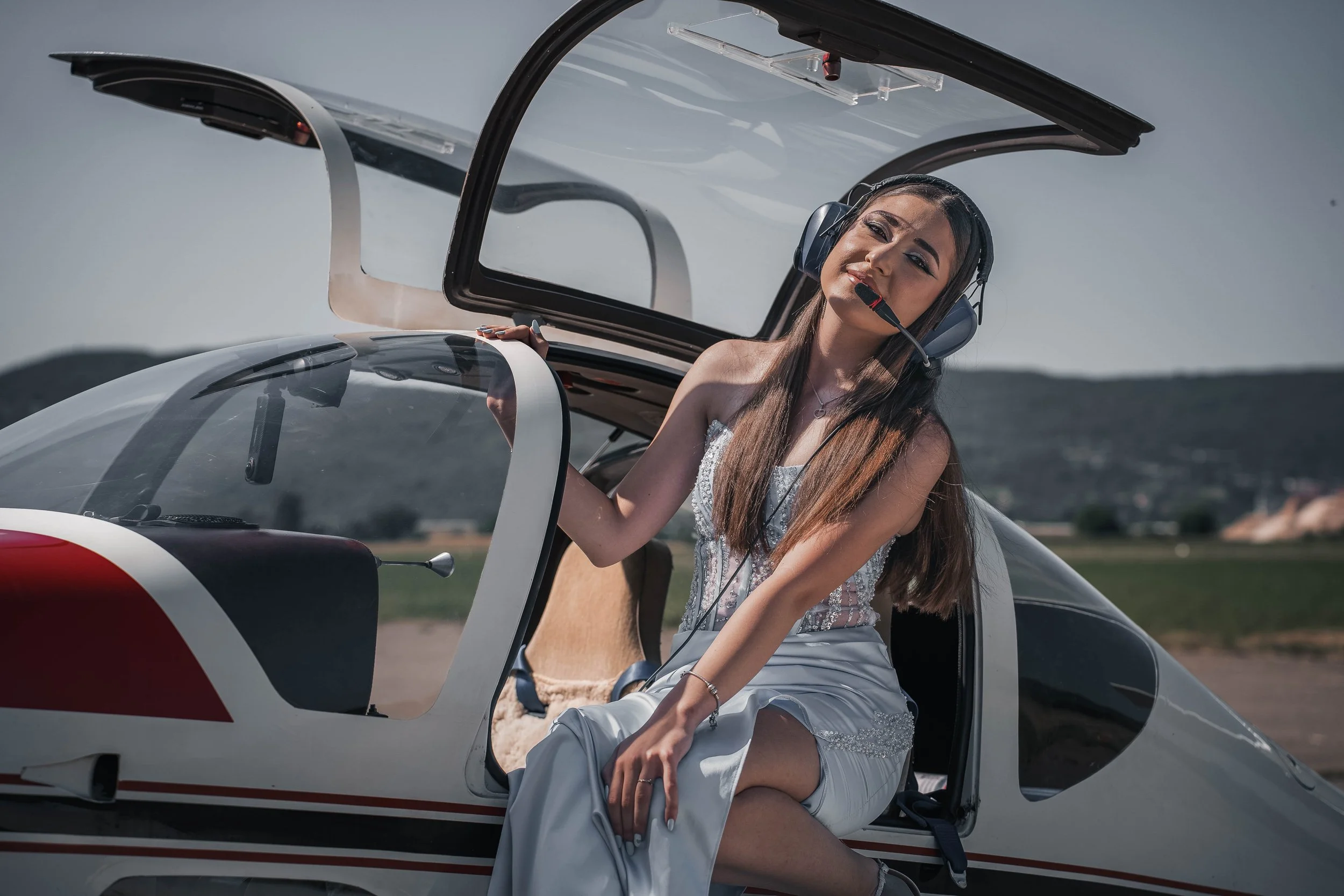 A woman with long brown hair wearing a headset and white clothing, sitting inside an open cockpit of a small airplane, with her arm resting on the edge of the aircraft.