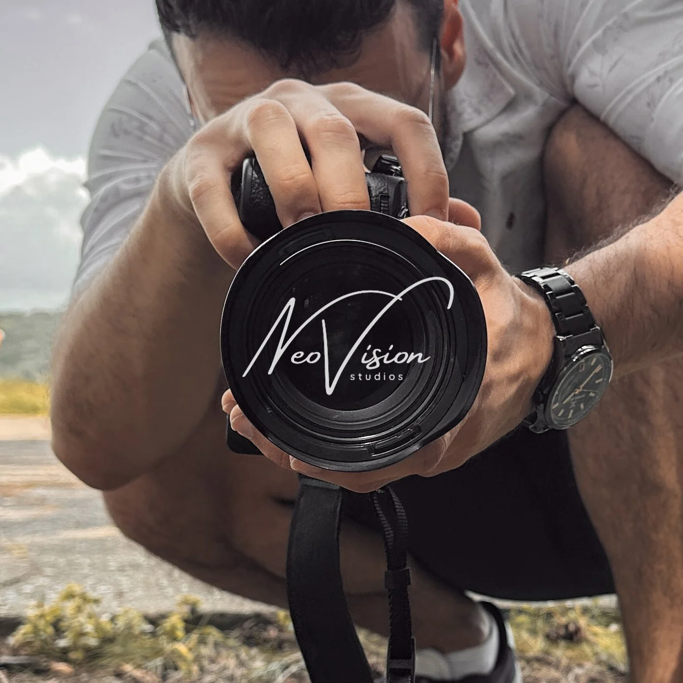A person with short dark hair wearing a white shirt and a wristwatch, holding a camera close to the lens with the logo 'Neo Vision Studios' displayed on it. The person appears to be outdoors, possibly near a body of water.