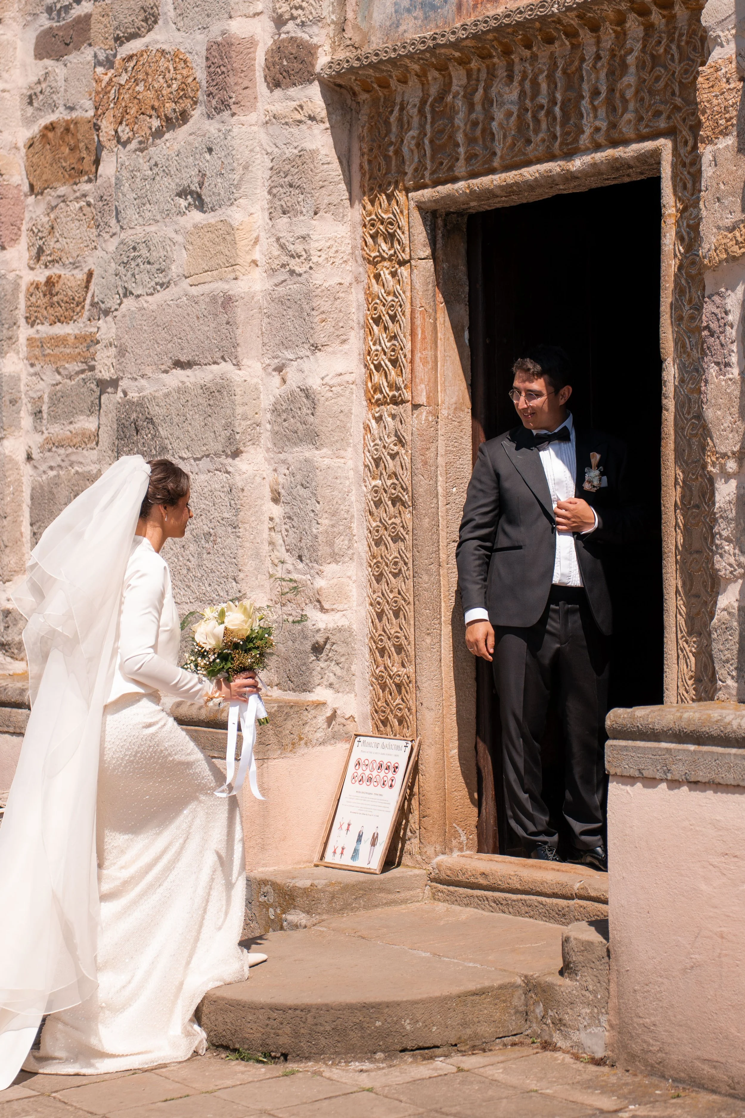 A wedding scene outside a stone building with ornate carved window frames. A bride in a white gown and veil holds a bouquet, greeting a groom in a black tuxedo and glasses who is smiling at her.