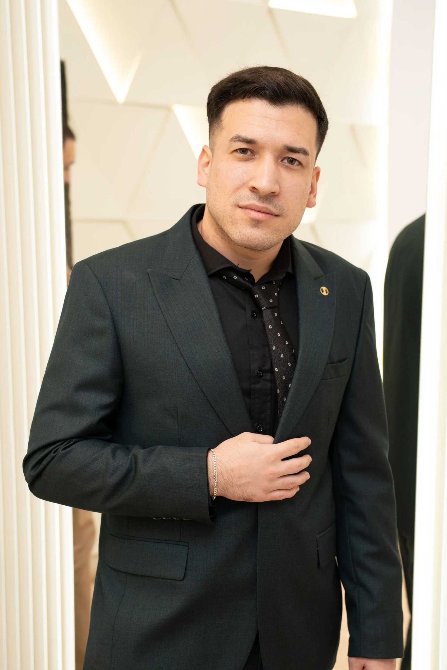 A man in a dark suit and black shirt, standing indoors with a neutral expression, against a light-colored background with geometric patterns.