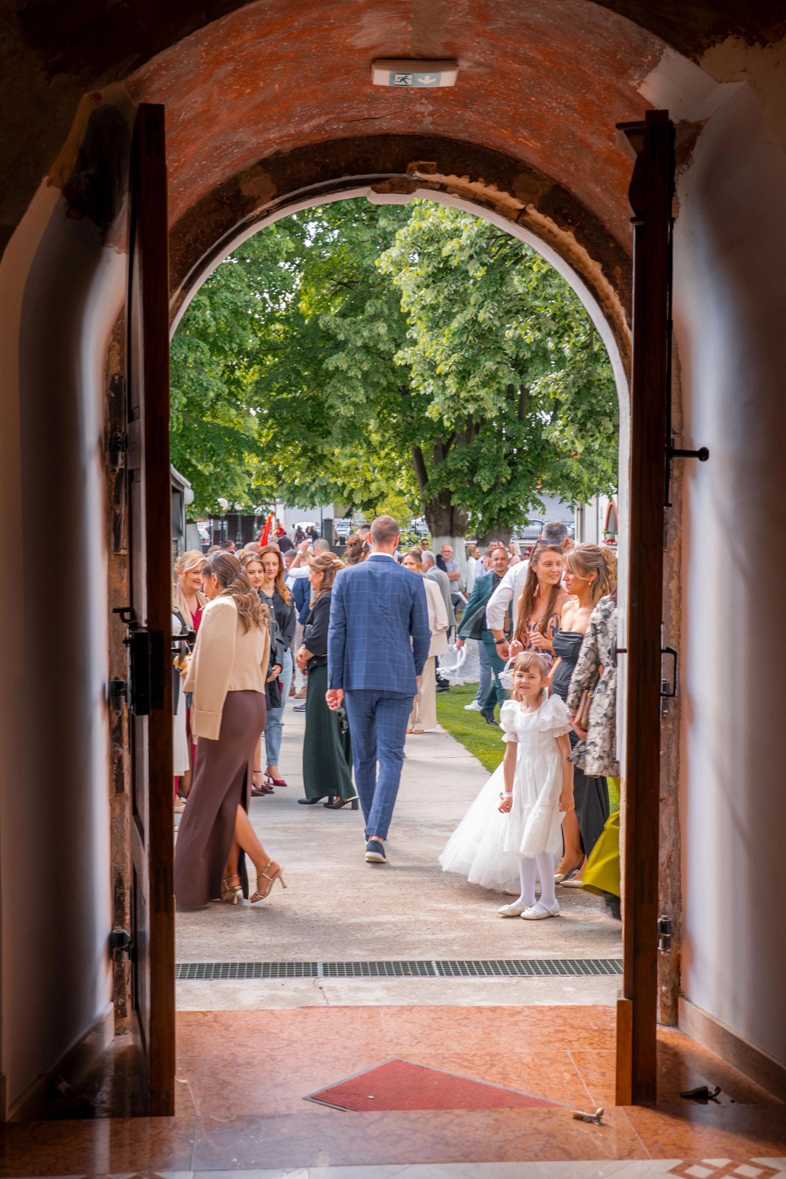 People gathered outdoors for a wedding or celebration, seen through a large open doorway with brick archway and greenery in the background.