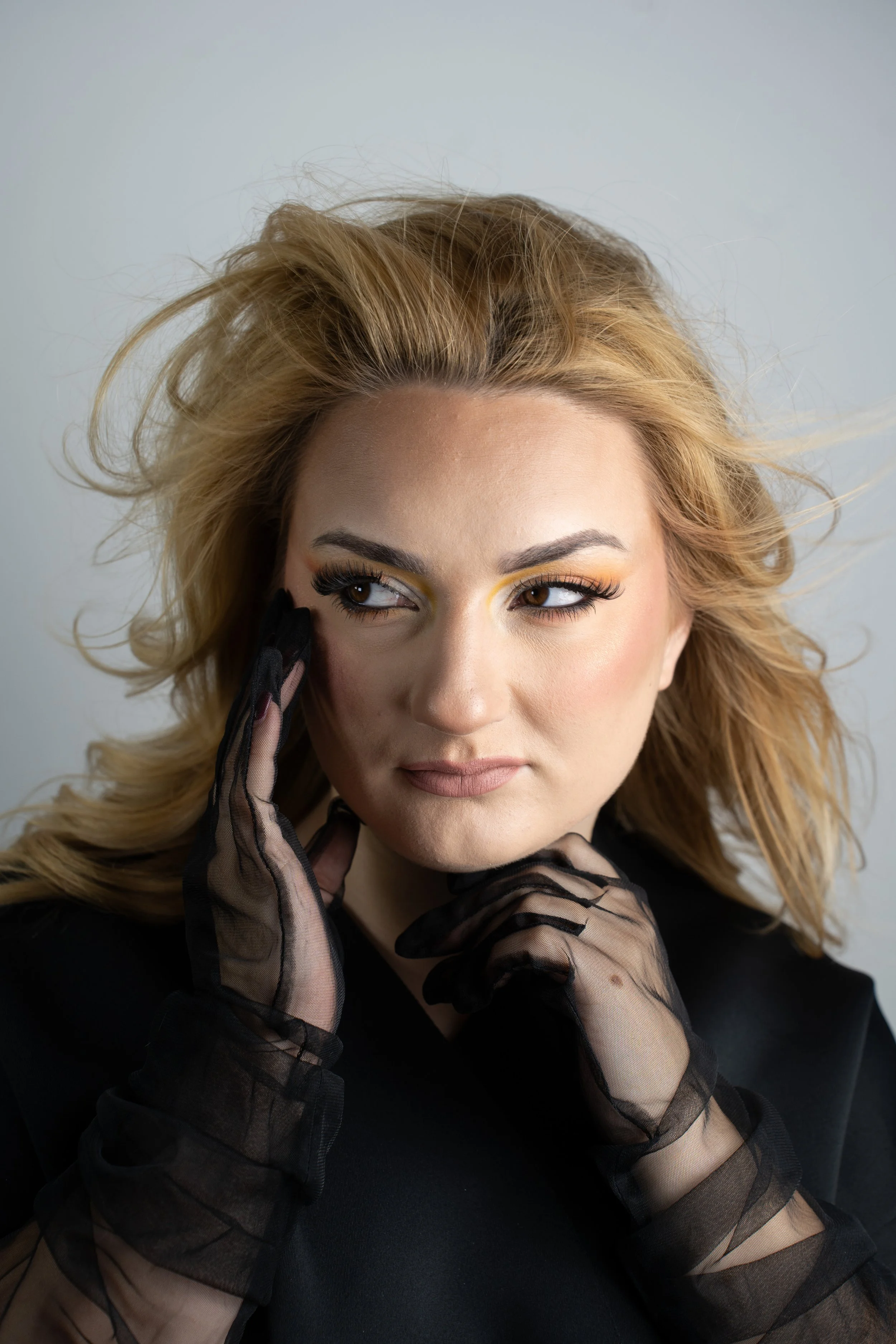 Close-up of a woman with blonde curly hair, wearing black sheer gloves, and makeup with yellow eyeshadow, looking sideways with her fingers near her face.