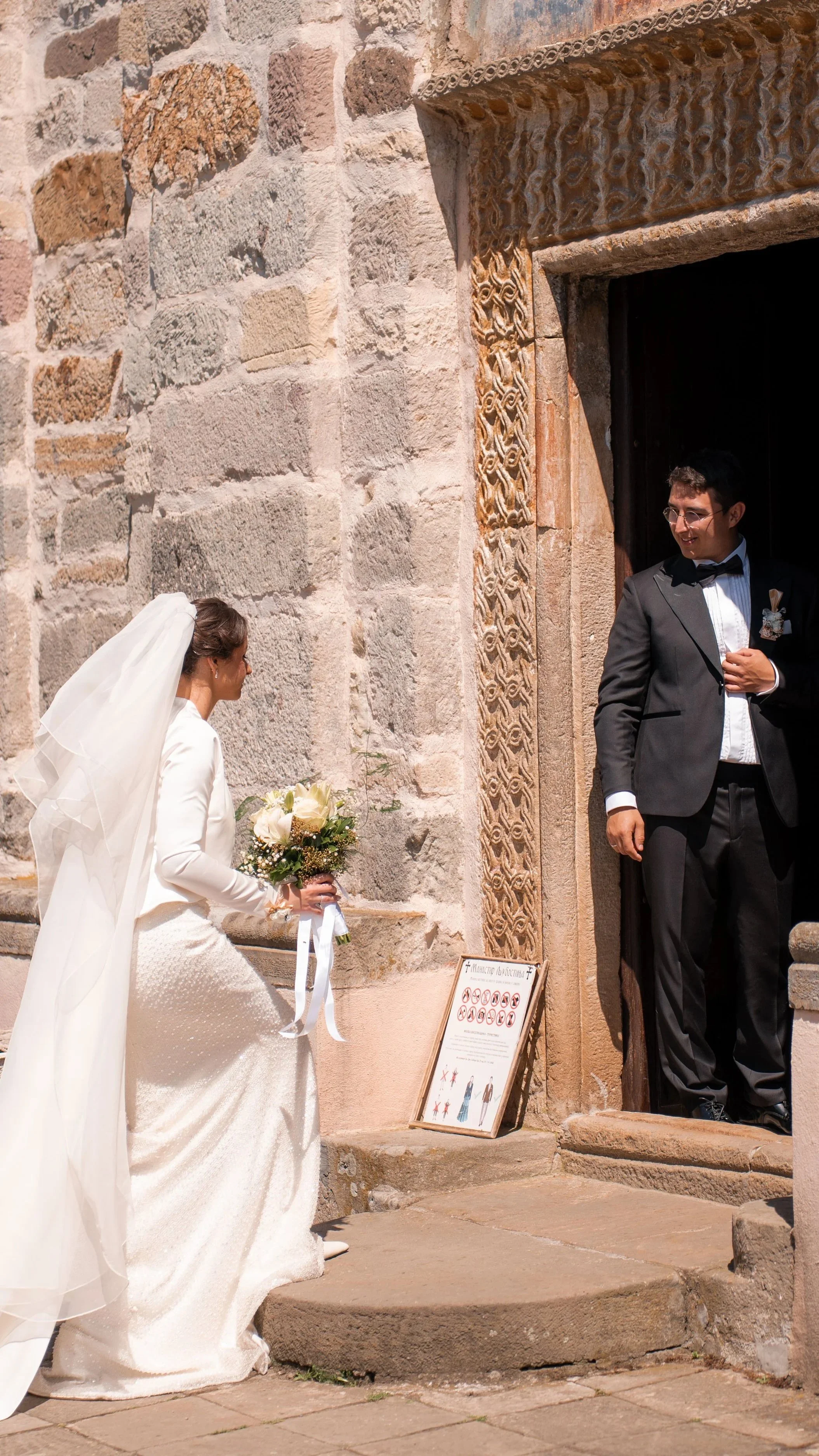 A bride in a white gown kneels holding a bouquet of white roses, facing a groom in a black tuxedo, outside a historic stone building during a wedding ceremony.