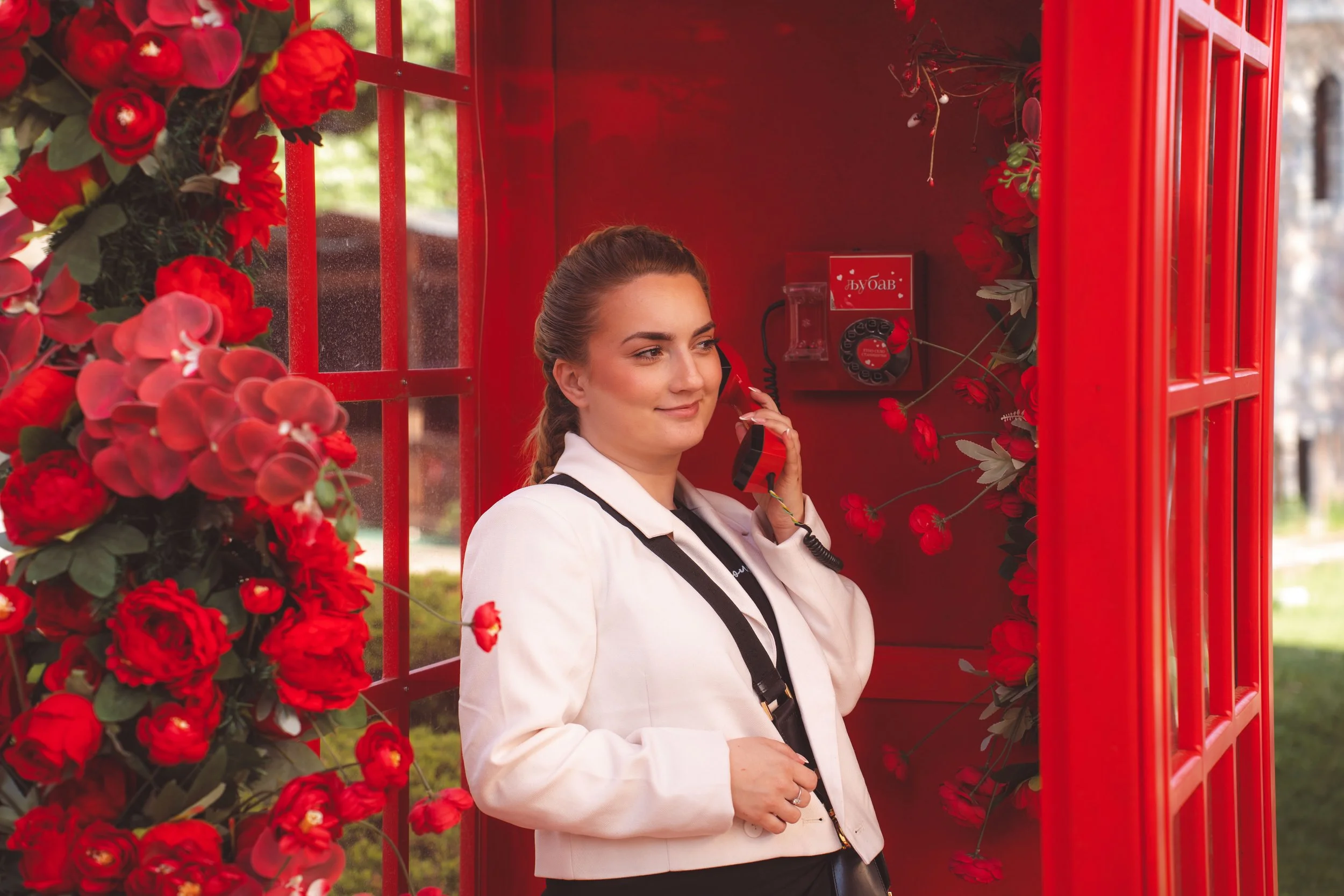 A woman in a white jacket is talking on a red telephone inside a red booth decorated with red flowers.