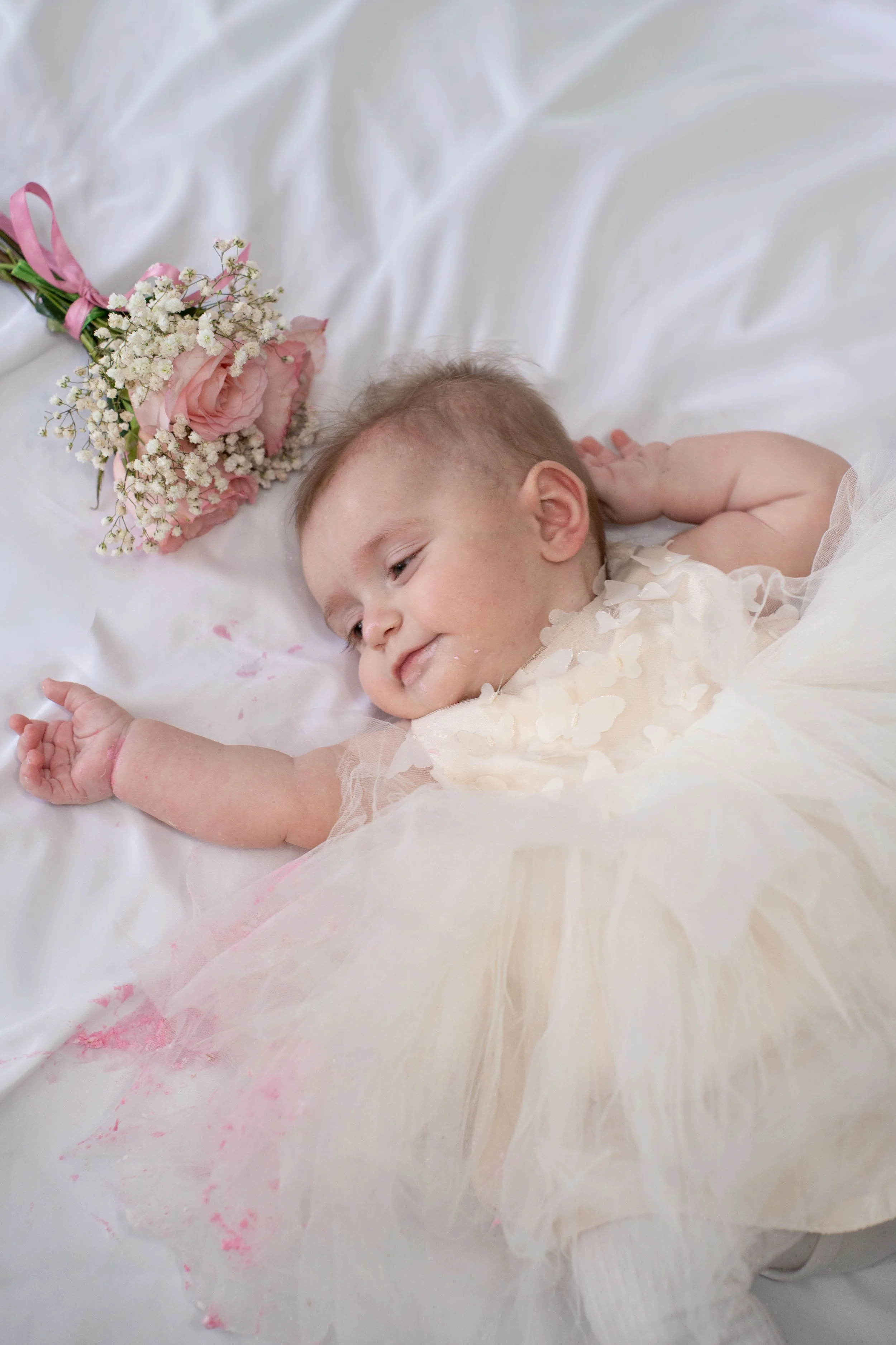 A baby girl lying on a white sheet with a pink and white bouquet of flowers beside her. She is dressed in a light-colored dress with floral details and appears to be sleeping or resting.