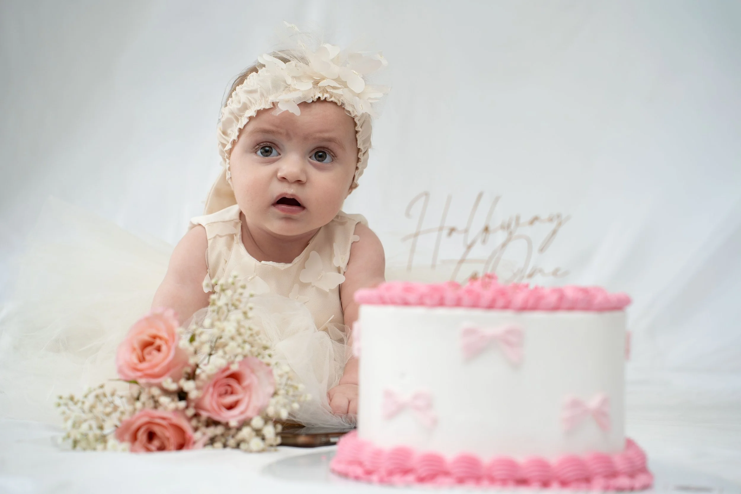 A baby girl with blue eyes and a floral headband, dressed in light-colored clothing, sits in front of a birthday cake with pink icing and a pink bow decoration, surrounded by pink roses and white flowers, with a white background and a faint birthday message.