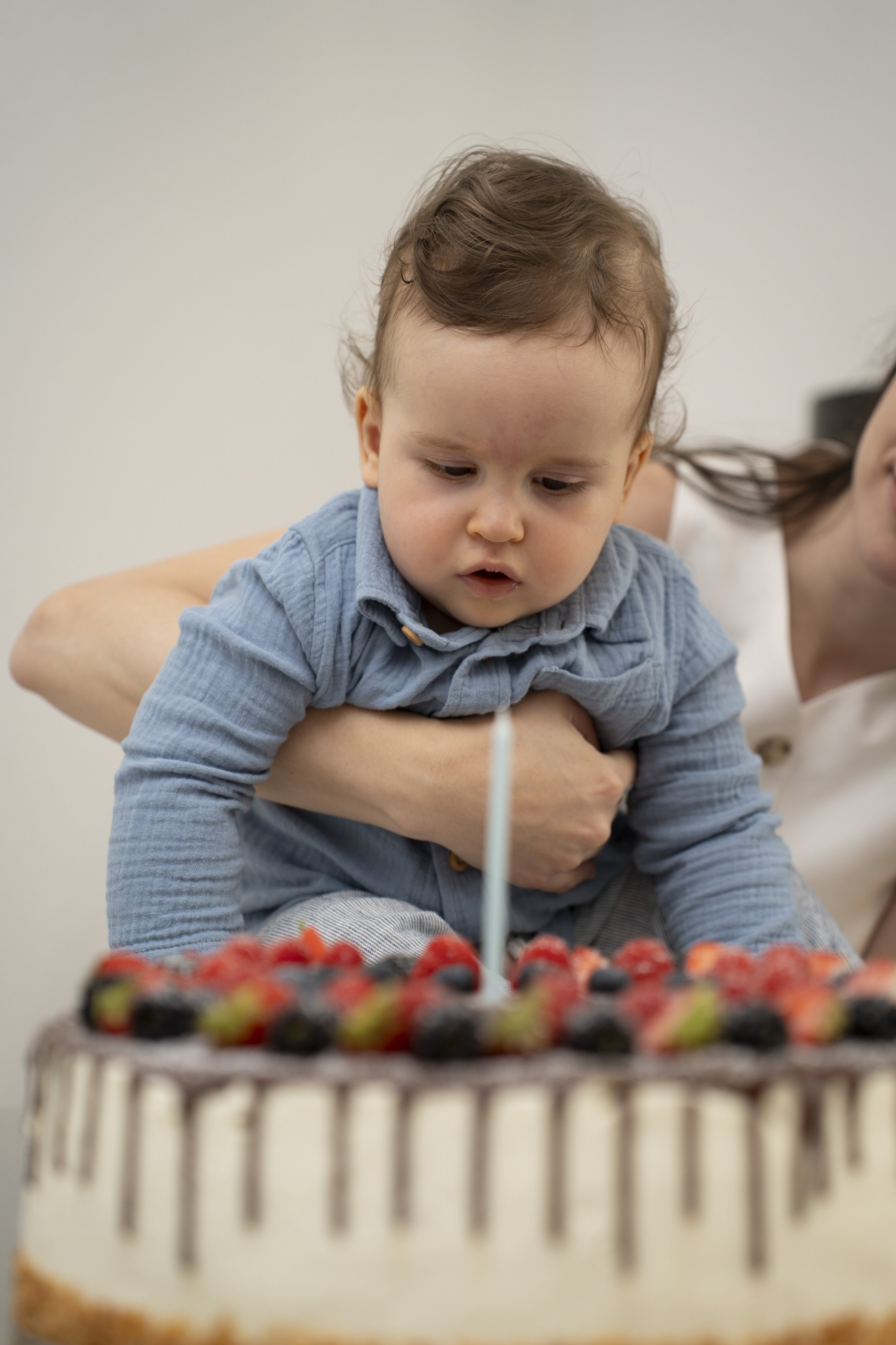 A young boy with brown hair and a blue shirt leaning over a birthday cake with a single candle, surrounded by strawberries and blueberries.